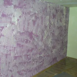 Fioletowa ściana z dekoracyjną fakturą w paski i łuki, kontrastująca z gładką, jasnozieloną ścianą w rogu pomieszczenia, drewniana podłoga w tle.