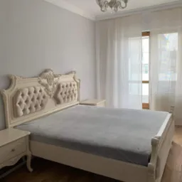 Elegancka sypialnia z łóżkiem w stylu Ludwika, z kremowym zagłówkiem obitym tkaniną w guziki, drewnianą podłogą i zwiewnymi firanami.