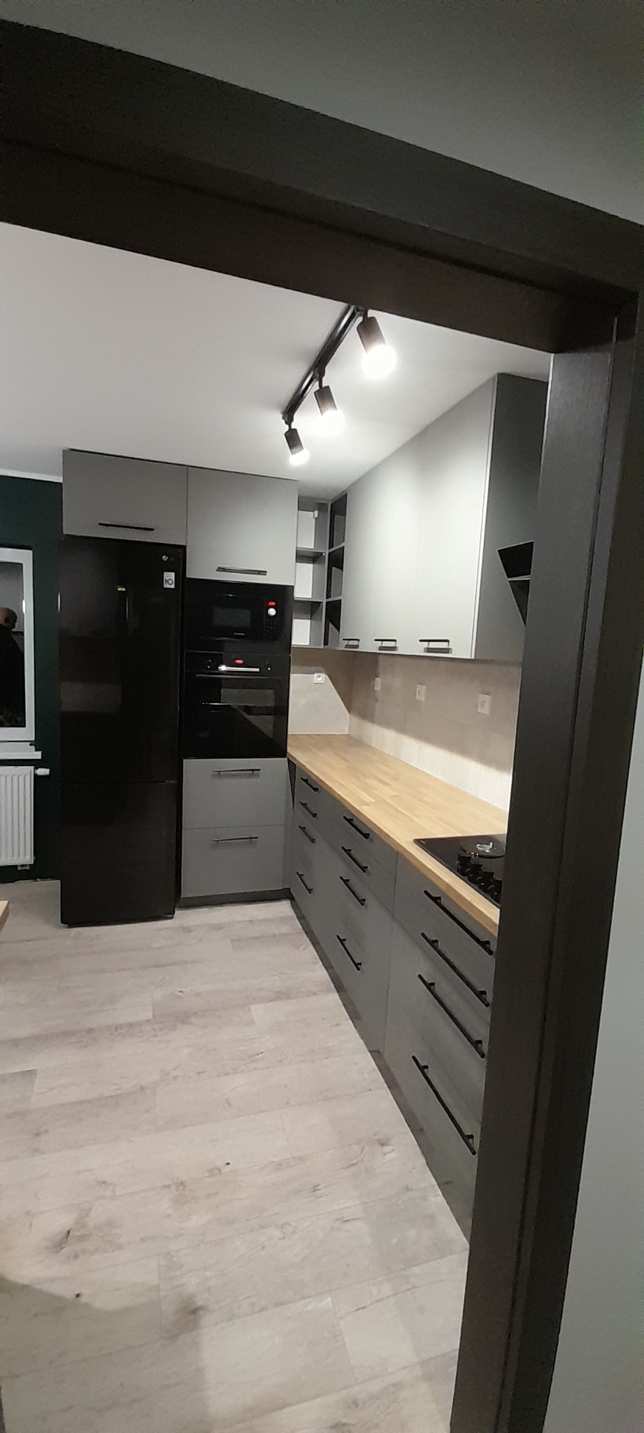 Szara kuchnia z drewnianym blatem i czarnymi uchwytami, oświetlona reflektorami, widok z perspektywy wejścia.