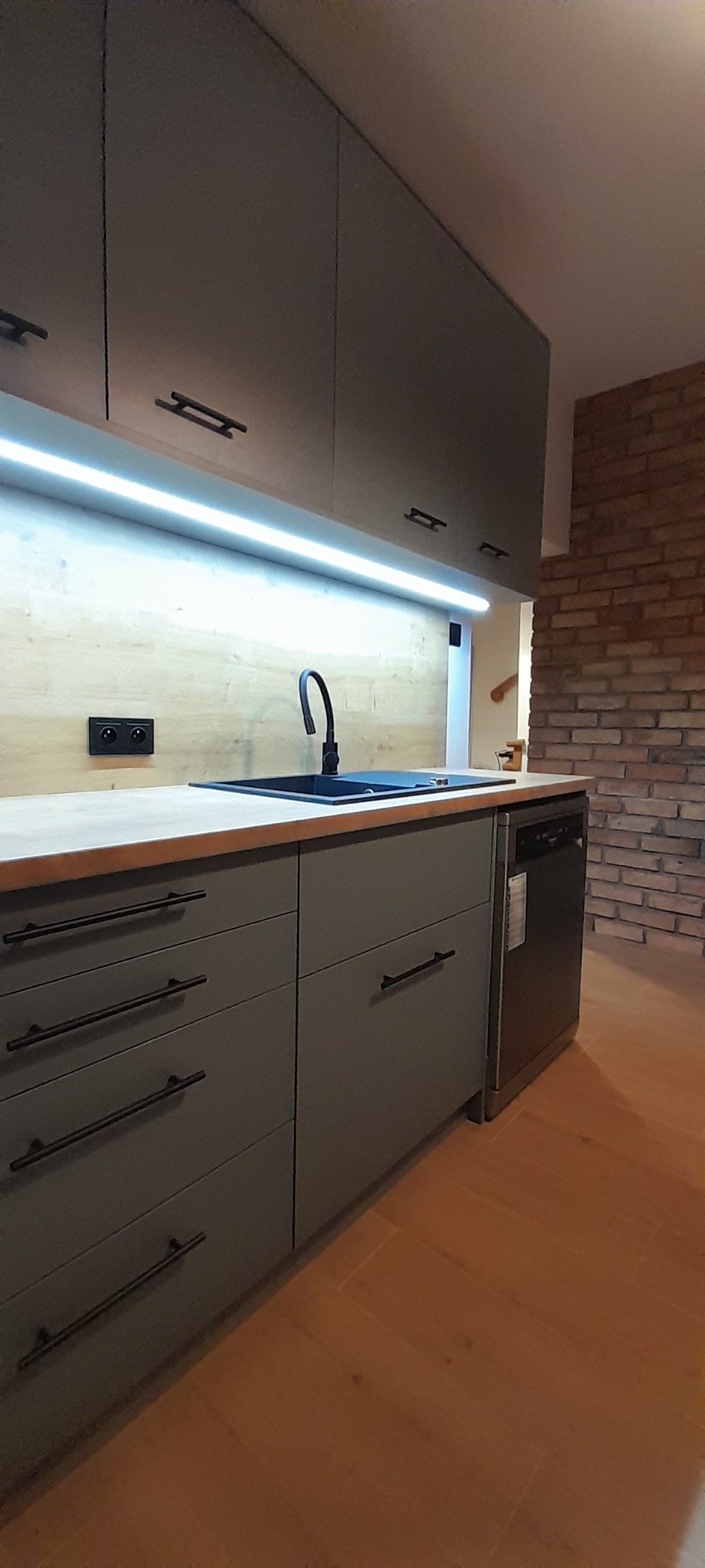 Szare szafki kuchenne z drewnianym blatem, czarnym zlewem i baterią, podświetlane listwą LED, z fragmentem ściany z cegły w tle.