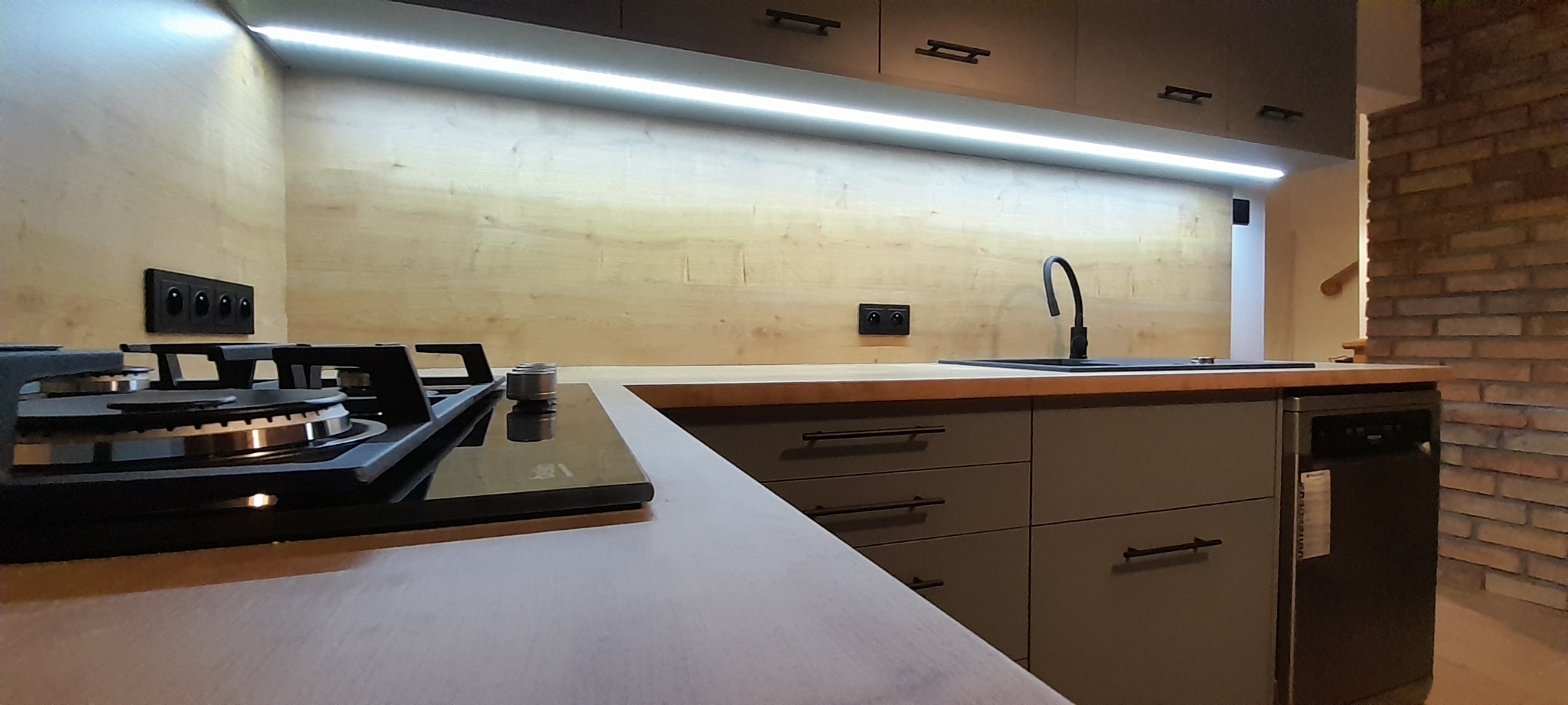 Nowoczesna kuchnia z frontami w odcieniach szarości, drewnianym blatem i oświetleniem LED podszafkowym, widoczna płyta indukcyjna i fragment ceglanej ściany w tle.