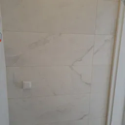 Ściana wyłożona jasnymi, marmurkowymi płytkami ceramicznymi, z widocznym włącznikiem światła i fragmentem białej ościeżnicy drzwi po prawej stronie.