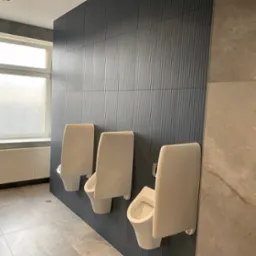 Trzy białe pisuary zamontowane na ciemnej, prążkowanej ścianie w nowoczesnej toalecie publicznej, z szarymi płytkami podłogowymi i fragmentem okna z widocznym grzejnikiem.