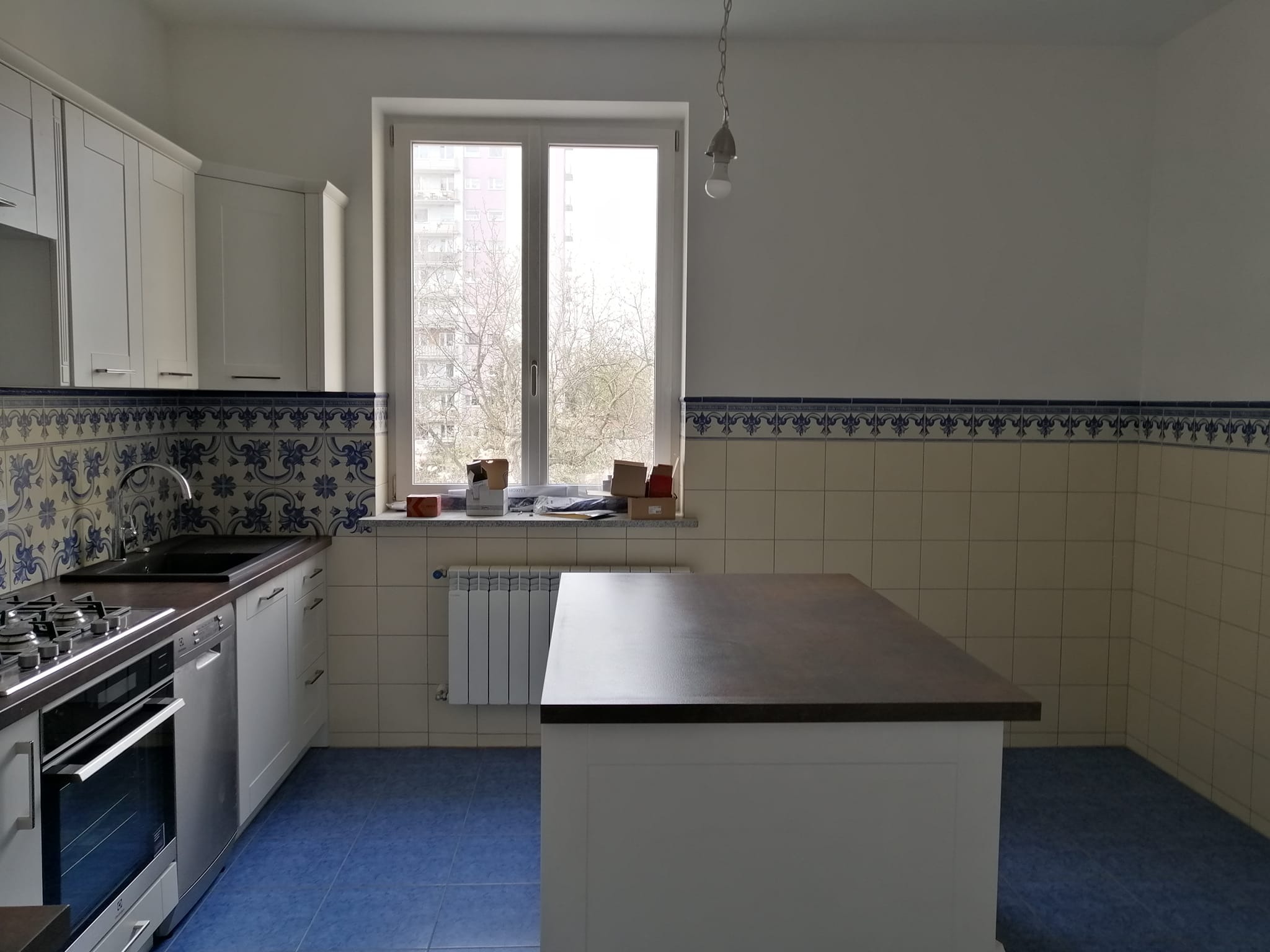 Jasna kuchnia z niebieską podłogą i wzorzystymi kaflami nad blatem, wyspa kuchenna z ciemnym blatem, okno z widokiem na budynek mieszkalny