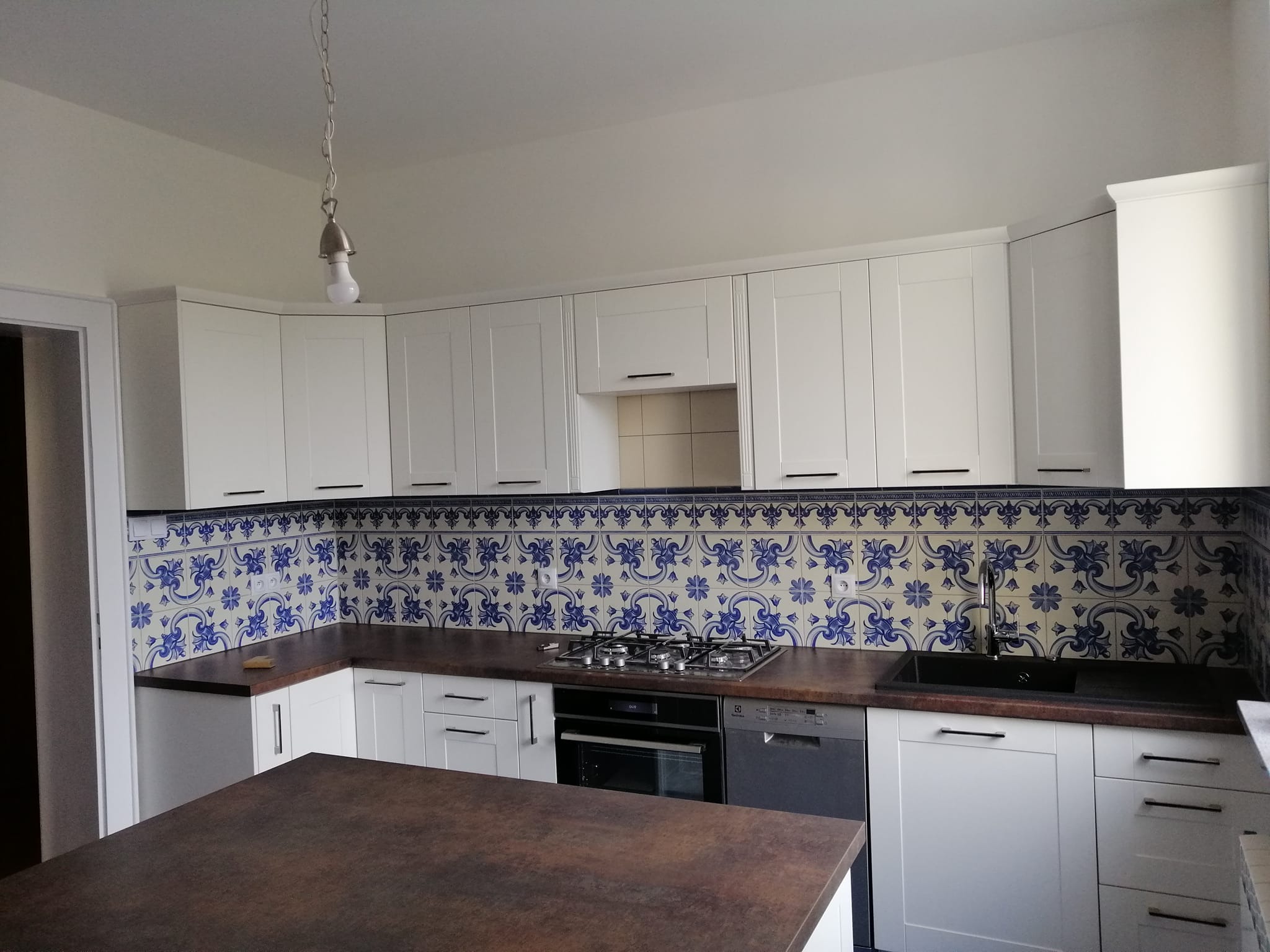 Nowoczesna kuchnia z białymi szafkami, brązowym blatem i dekoracyjnymi płytkami w stylu portugalskim nad blatem, oświetlona pojedynczą żarówką wiszącą na łańcuchu.