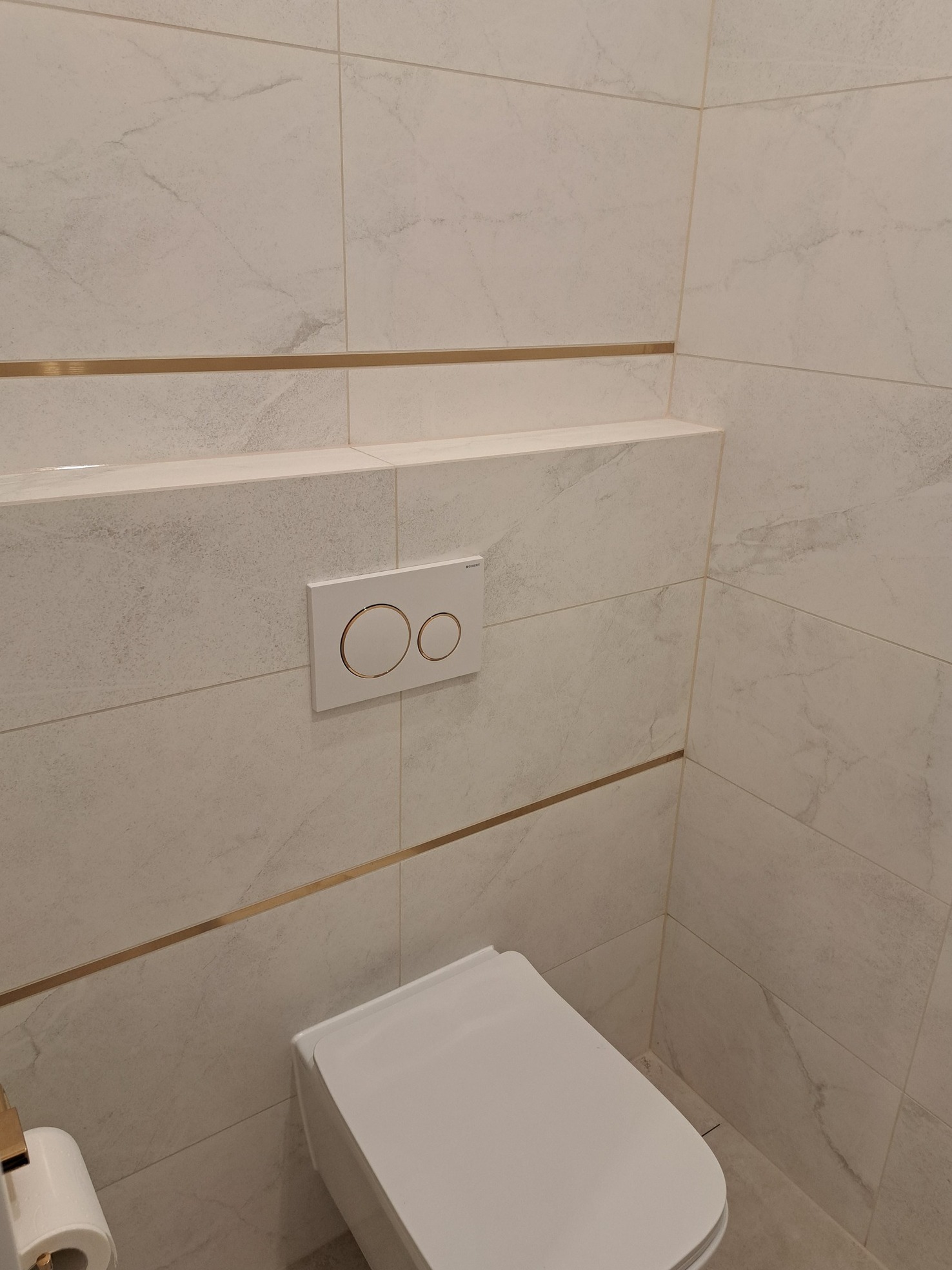 Nowoczesna toaleta wisząca z białymi płytkami imitującymi marmur, złotymi listwami dekoracyjnymi i podtynkowym systemem spłukiwania z dwoma przyciskami.
