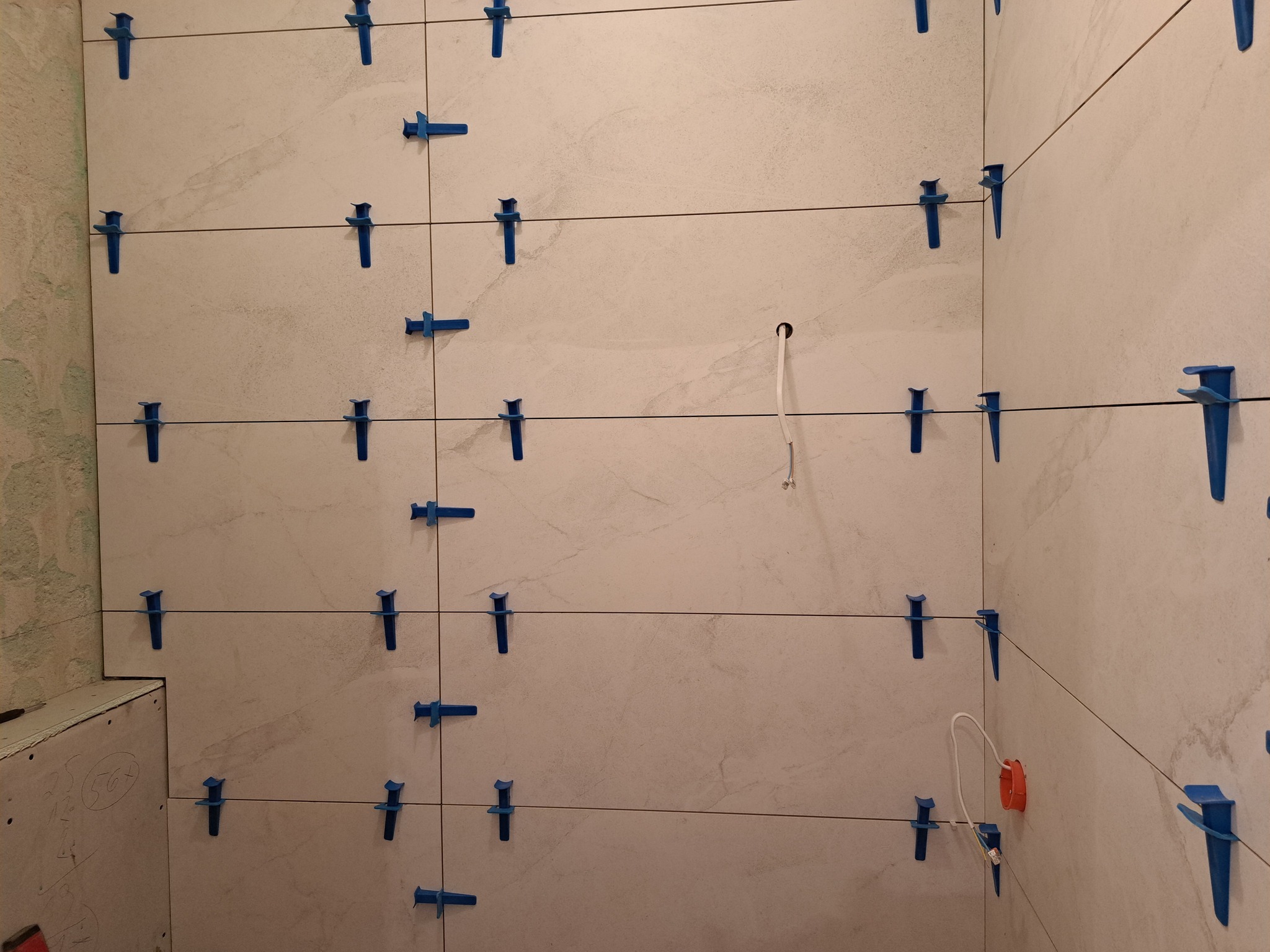 Ściana w trakcie układania płytek, jasne płytki imitujące kamień, niebieskie klipsy poziomujące, widoczne kable elektryczne i puszka, fragment ściany w stanie surowym.