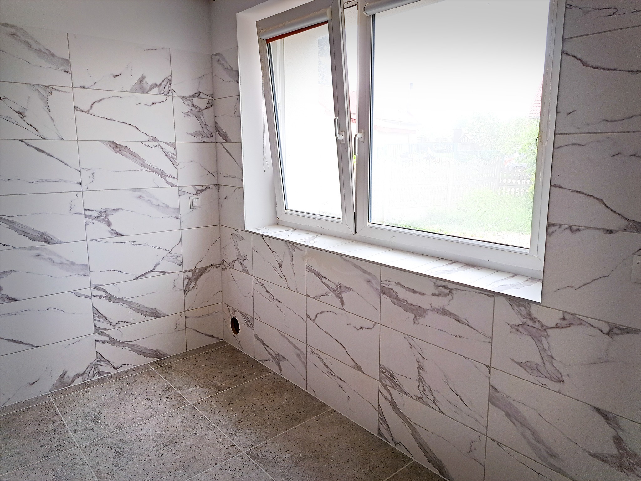 Świeżo wykafelkowana łazienka z oknem, płytki imitujące marmur na ścianach, szare płytki podłogowe, widok przez uchylone okno na zieleń.