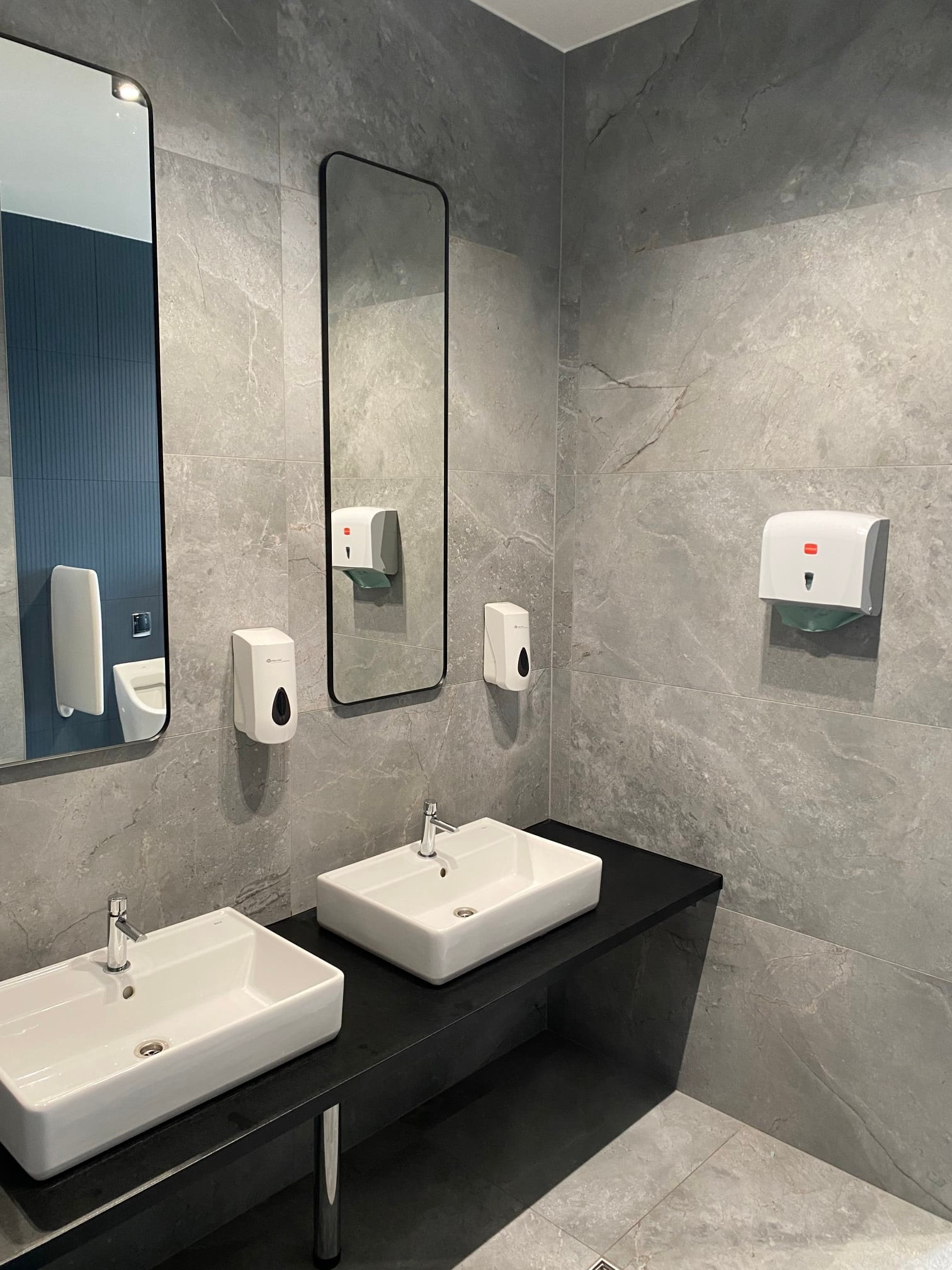 Nowoczesna łazienka publiczna z dwoma białymi umywalkami na czarnym blacie, szare płytki imitujące kamień na ścianach, dwa lustra w czarnych ramach, dozowniki mydła i ręczników papierowych.