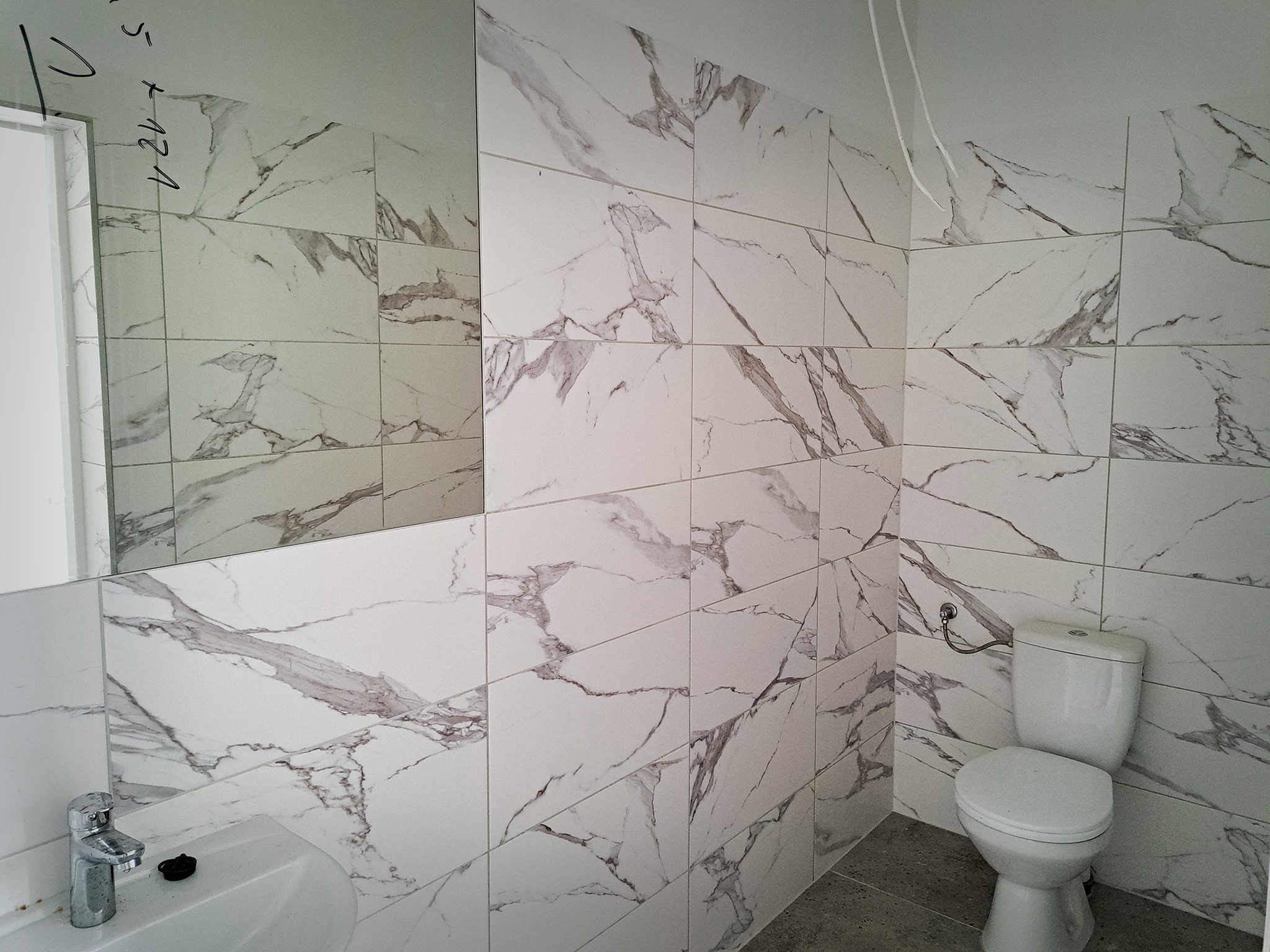 Nowoczesna łazienka z białymi płytkami imitującymi marmur, minimalistyczna umywalka z chromowaną baterią, podwieszana toaleta i duże lustro.