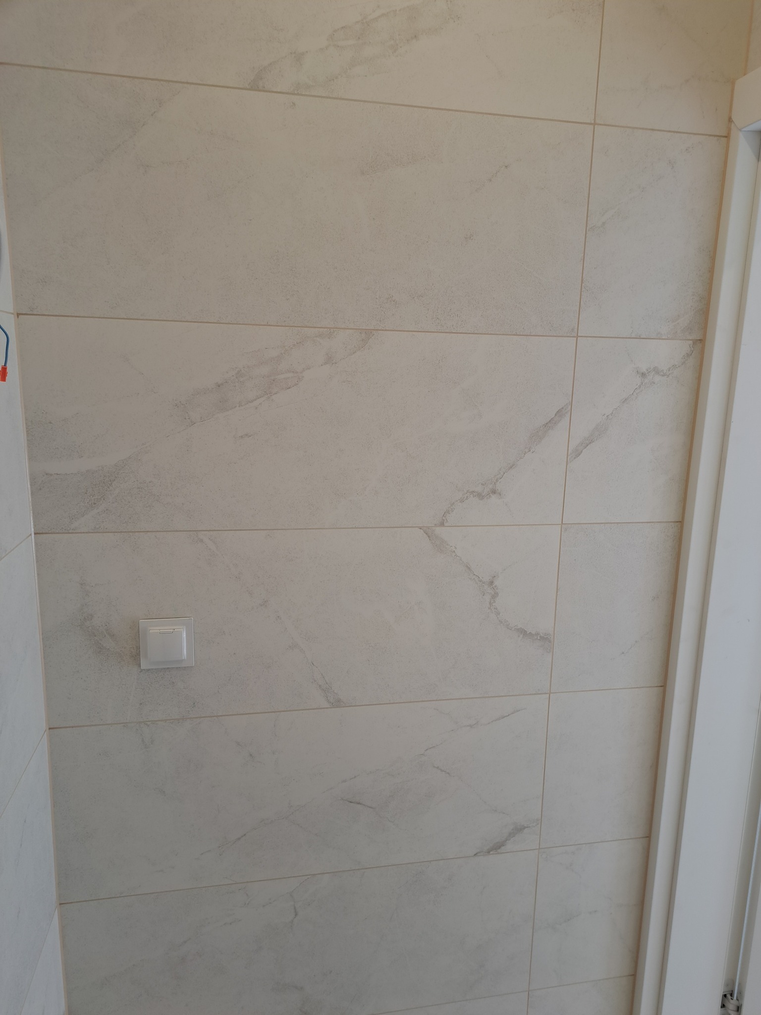 Ściana wyłożona jasnymi, marmurkowymi płytkami ceramicznymi, z widocznym włącznikiem światła i fragmentem białej ościeżnicy drzwi po prawej stronie.