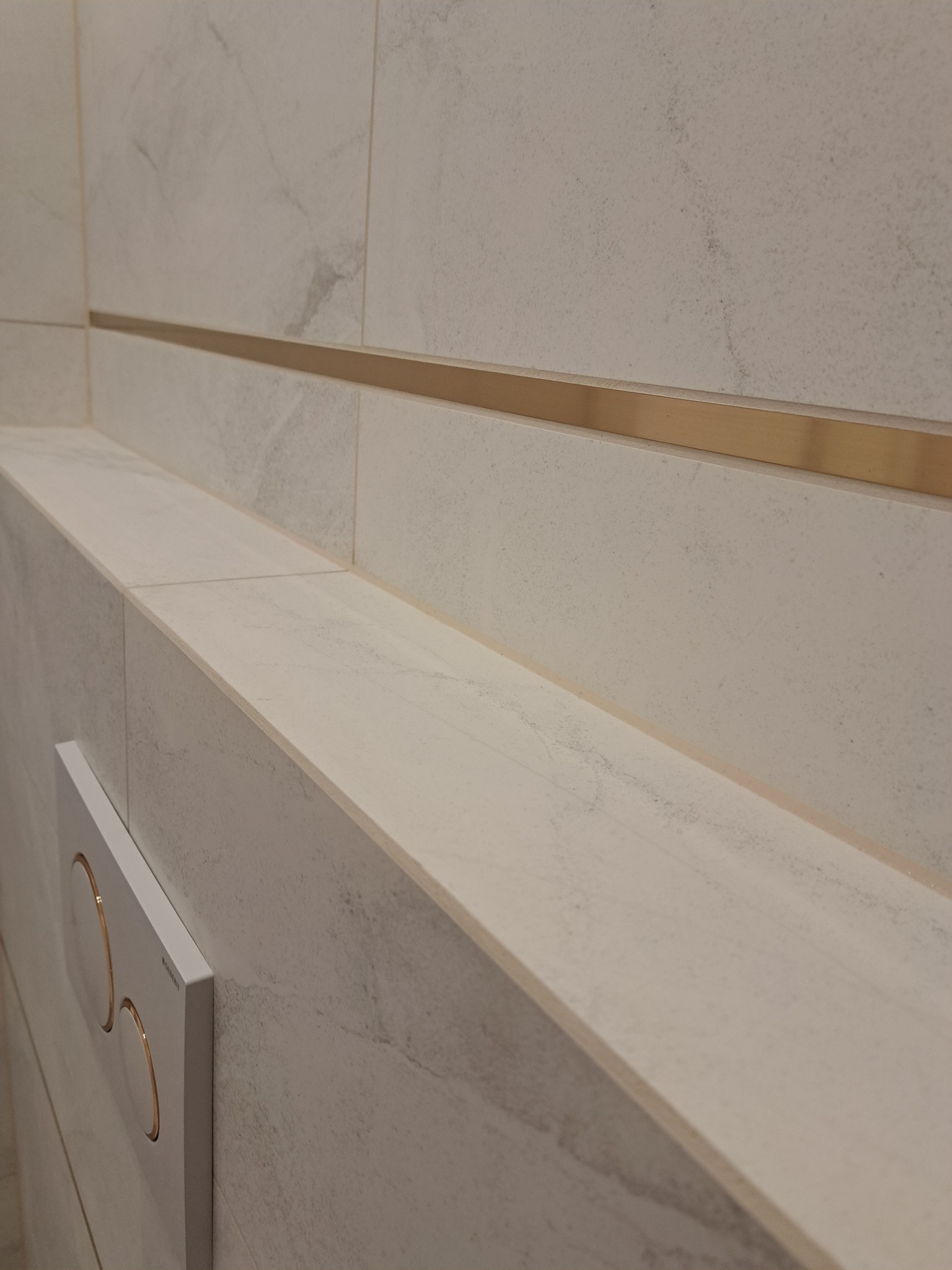 Beżowe płytki imitujące marmur z złotą listwą dekoracyjną i przyciskiem spłuczki w nowoczesnej łazience.