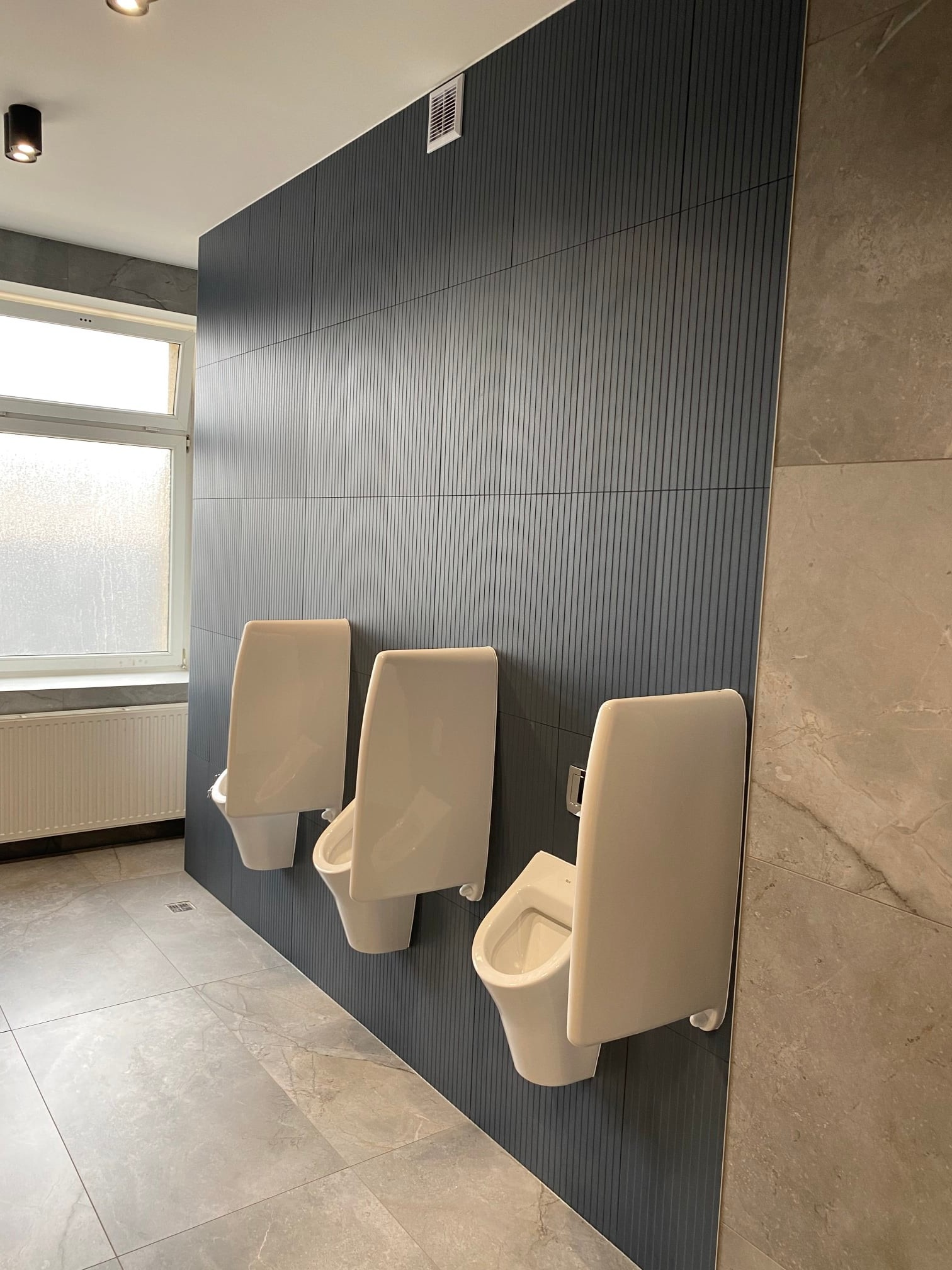 Nowoczesna toaleta publiczna z trzema białymi pisuarami zamontowanymi na ciemnoszarej ścianie z pionowymi panelami, uzupełniona płytkami imitującymi marmur i oświetlona punktowym światłem.
