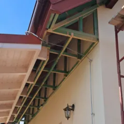 Konstrukcja z drewna przy elewacji budynku, prawdopodobnie przygotowanie pod ocieplenie. Widoczne lampy zewnętrzne i fragment rusztowania.