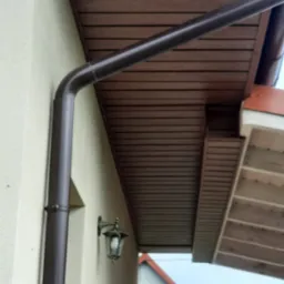 Brązowa rynna pionowa na elewacji domu z beżową ścianą i drewnianym podbitkiem. Widoczna lampa elewacyjna i fragment dachu sąsiedniego budynku.