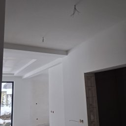 LUGIN-BUD Zbigniew Ługin - Wnętrze w trakcie wykańczania: białe ściany z gładzią, sufit z dekoracyjnymi elementami, widoczne kable i puszki elektryczne, okno z widokiem na zimowy krajobraz.