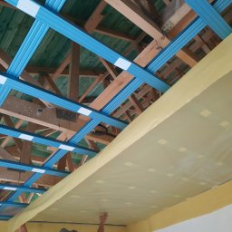 LUGIN-BUD Zbigniew Ługin - Montaż sufitu podwieszanego: niebieska konstrukcja z profili, drewniane elementy więźby dachowej i żółta folia paroizolacyjna. Widok z dołu na sufit w trakcie prac.