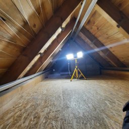 LUGIN-BUD Zbigniew Ługin - Poddasze z drewnianą konstrukcją i metalowymi profilami, oświetlone lampą na trójnogu. Widoczna podłoga z płyt OSB i elementy mocujące do izolacji.