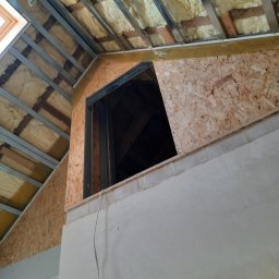 LUGIN-BUD Zbigniew Ługin - Skośne poddasze z izolacją, widoczne płyty OSB i metalowy stelaż. Fragment ściany z nałożoną gładzią. Okno dachowe w górnym rogu kadru.