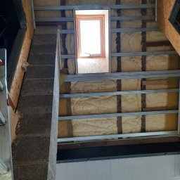 LUGIN-BUD Zbigniew Ługin - Poddasze z oknem dachowym w trakcie ocieplania wełną mineralną i montażu stelaża pod płyty G-K w Giżycku. Widoczny komin i fragment ściany z płyt.