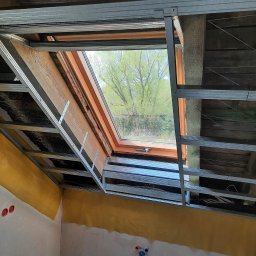 LUGIN-BUD Zbigniew Ługin - Montaż stelaża pod płytę karton-gips wokół okna dachowego na poddaszu. Widoczne drewniane elementy konstrukcyjne i fragment ściany z instalacją elektryczną i wodną.