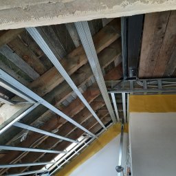 LUGIN-BUD Zbigniew Ługin - Skośny sufit poddasza z drewnianą konstrukcją, metalowym stelażem i żółtą izolacją, przygotowany do montażu płyt. Widoczne kable i fragment okna dachowego.