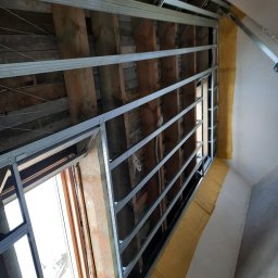 LUGIN-BUD Zbigniew Ługin - Szkielet metalowy pod zabudowę poddasza z widocznymi drewnianymi belkami stropowymi i oknem dachowym. Żółta taśma uszczelniająca na styku ściany i konstrukcji.