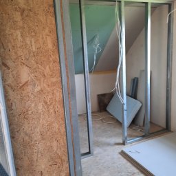 LUGIN-BUD Zbigniew Ługin - Szkielet ścianki działowej z metalowych profili, częściowo obłożony płytą OSB. Widoczne przewody elektryczne i płyty gipsowe na podłodze. Poddasze w tle.