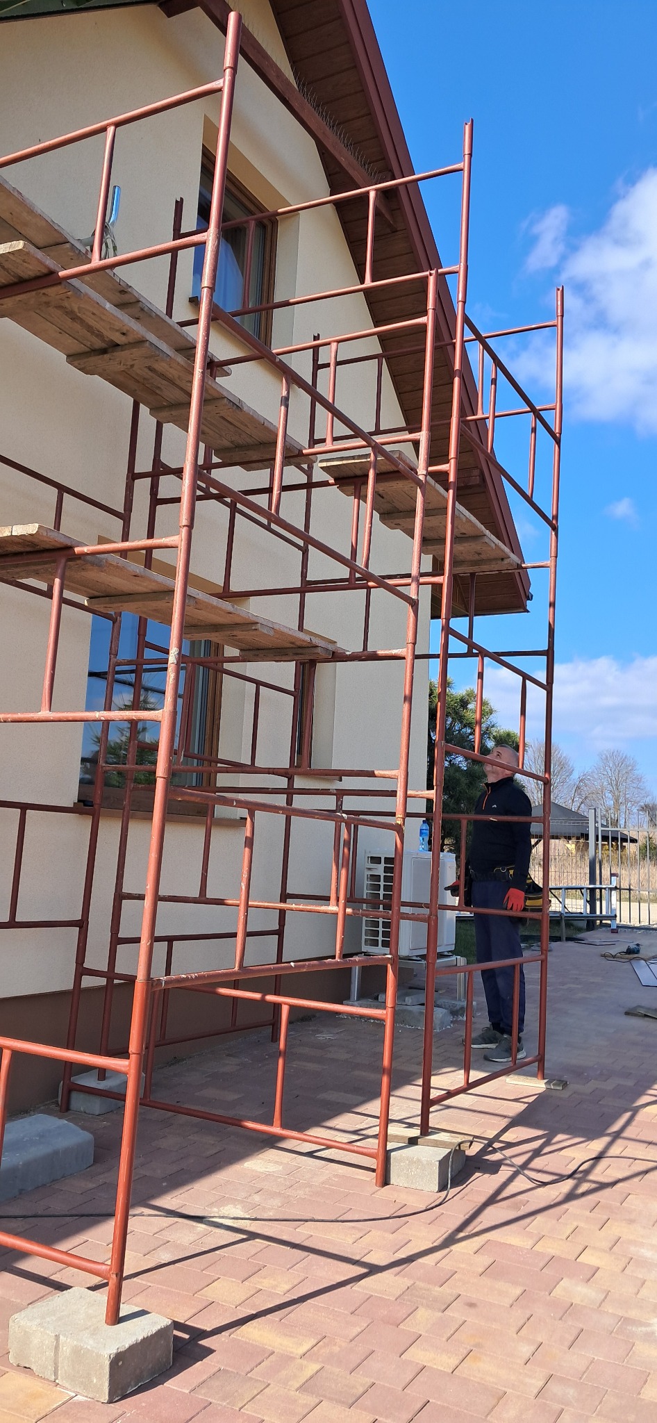 Dom w rusztowaniach, przygotowany do prac elewacyjnych. Widoczny pracownik w oddali, zarys błękitnego nieba w tle. Ujęcie z dołu, podkreślające wysokość konstrukcji.
