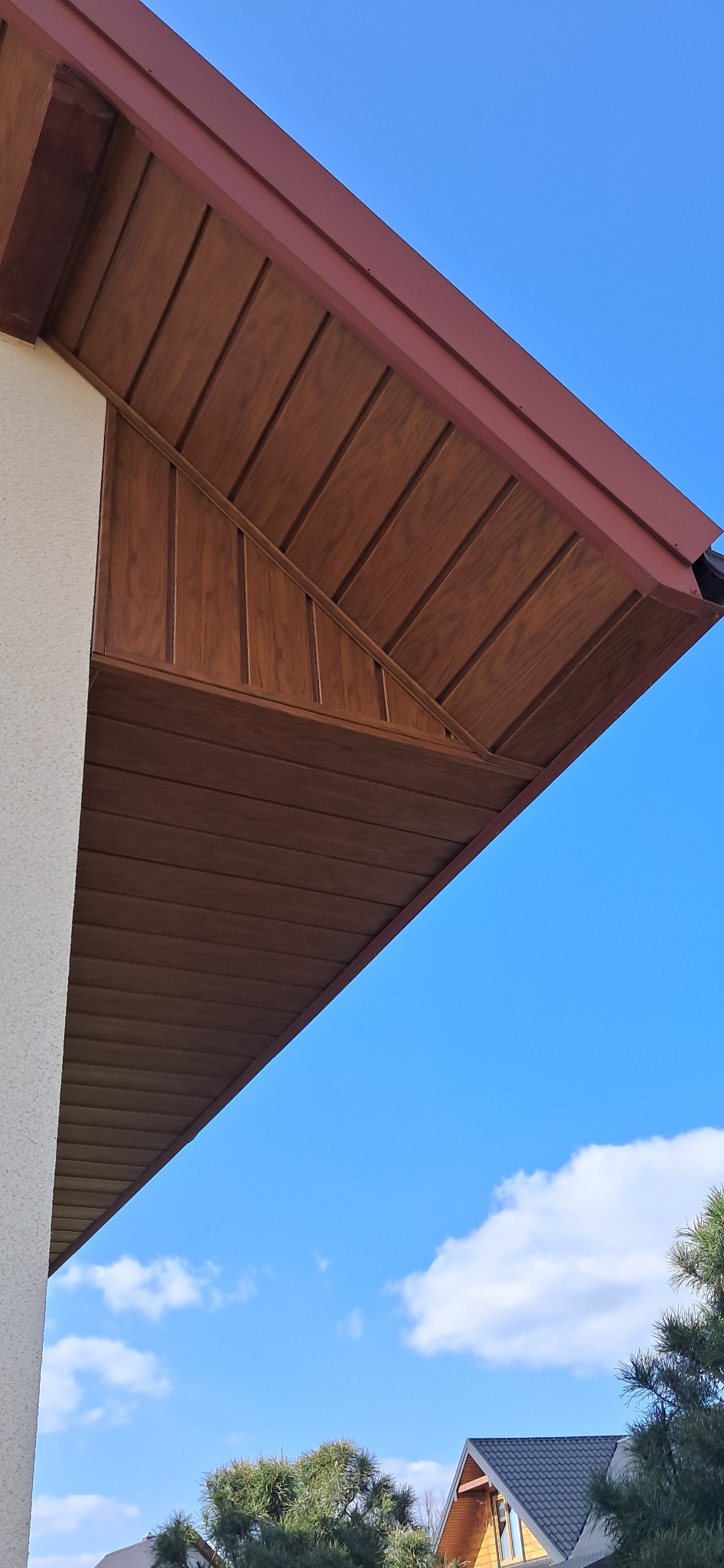 Drewniana podbitka dachowa w brązowym odcieniu, widoczna z dołu pod kątem, na tle błękitnego nieba z delikatnymi obłokami. Element wykończenia elewacji.