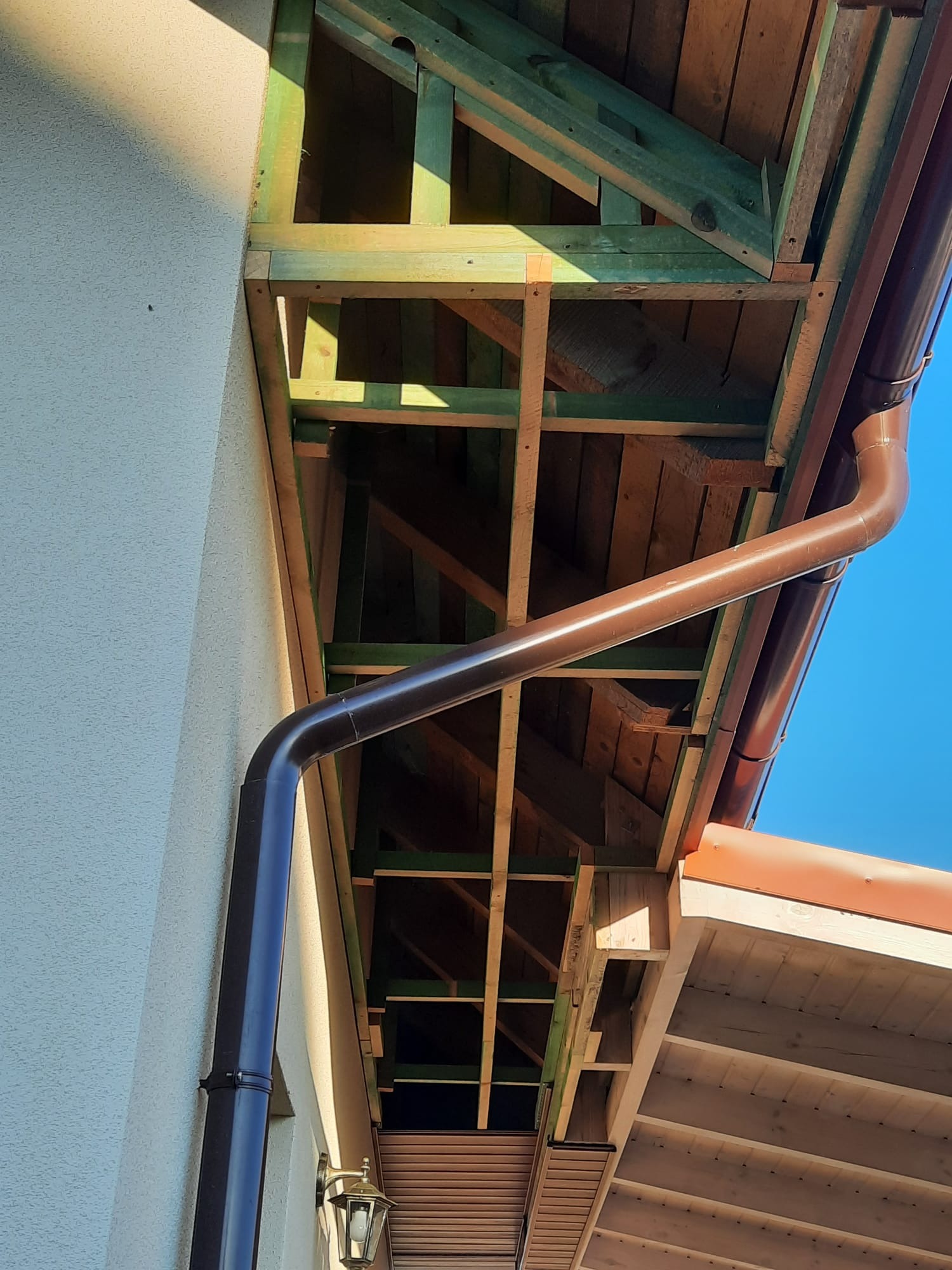 Konstrukcja drewniana okapu dachowego w trakcie montażu, z widoczną rynną w brązowym kolorze, na tle jasnej elewacji budynku i błękitnego nieba.