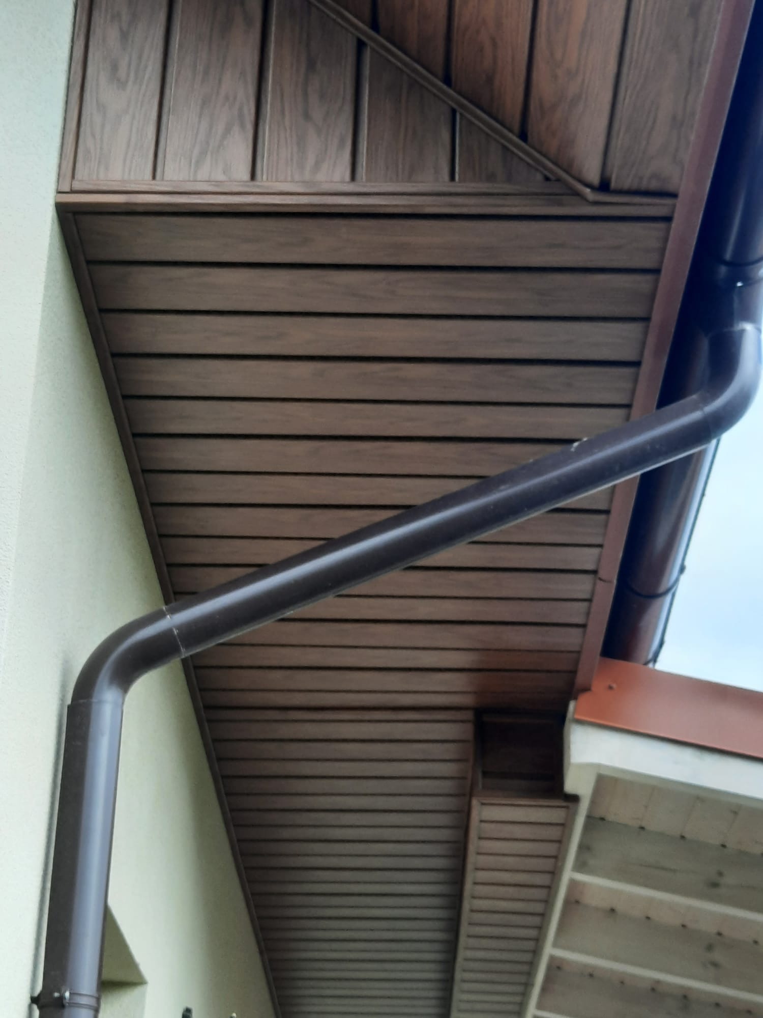 Brązowa rynna spustowa na tle drewnianej podbitki dachowej i jasnej ściany budynku. Widoczne poziome panele podbitki i fragment dachu.