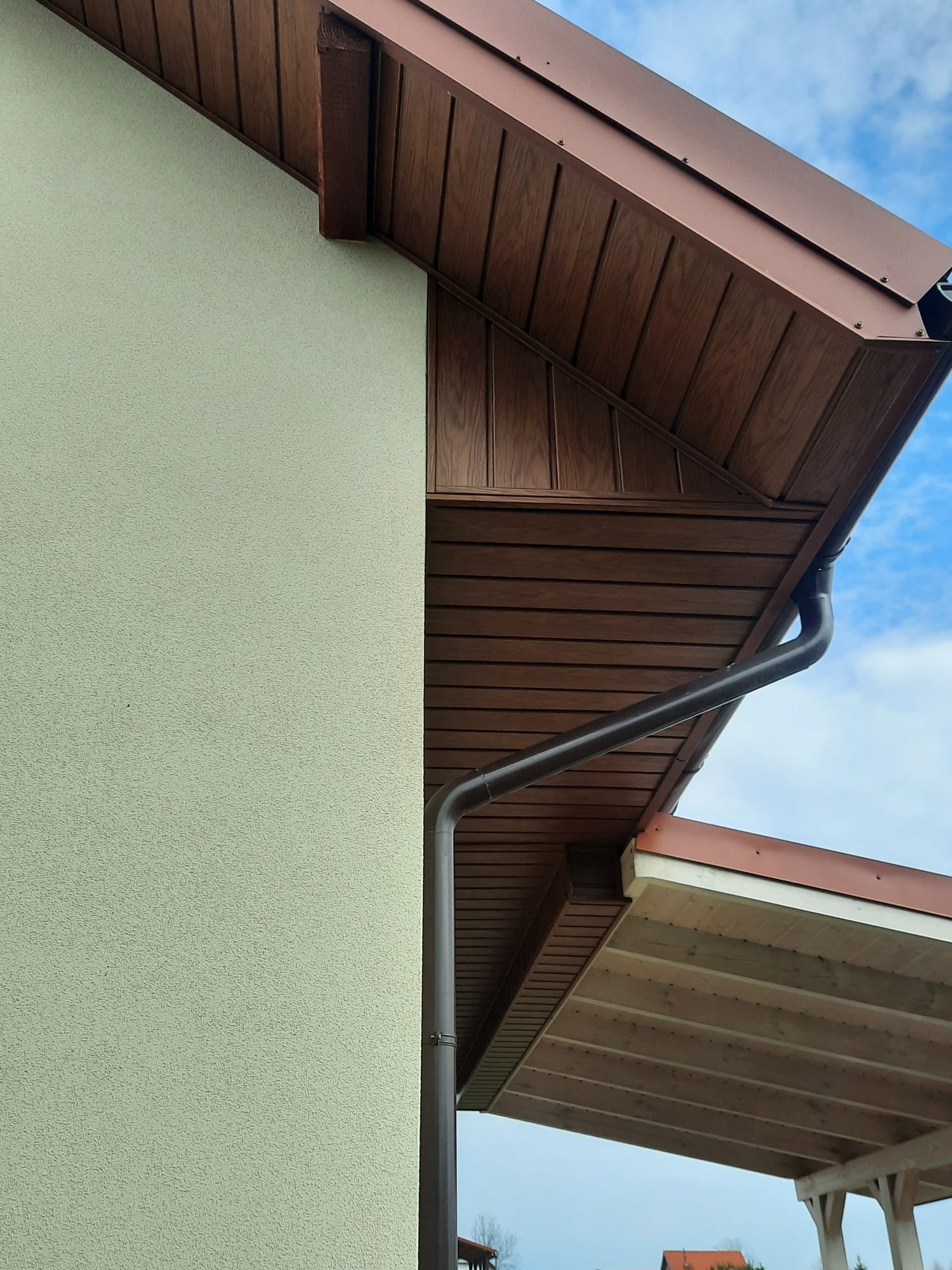 Fragment elewacji budynku z widocznym orynnowaniem i podbitką w brązowym odcieniu, na tle błękitnego nieba. Ściana w jasnym kolorze z wyraźną fakturą.