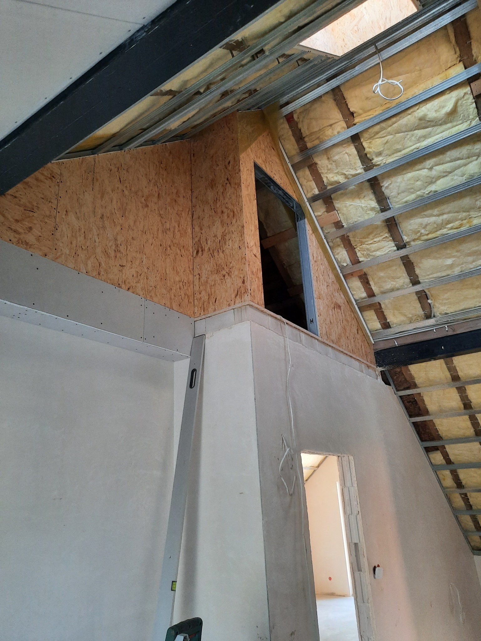 Skośne poddasze w trakcie prac: izolacja wełną mineralną, płyty OSB, metalowa konstrukcja, ściany po szpachlowaniu, poziomica oparta o ścianę. Widok z dołu.