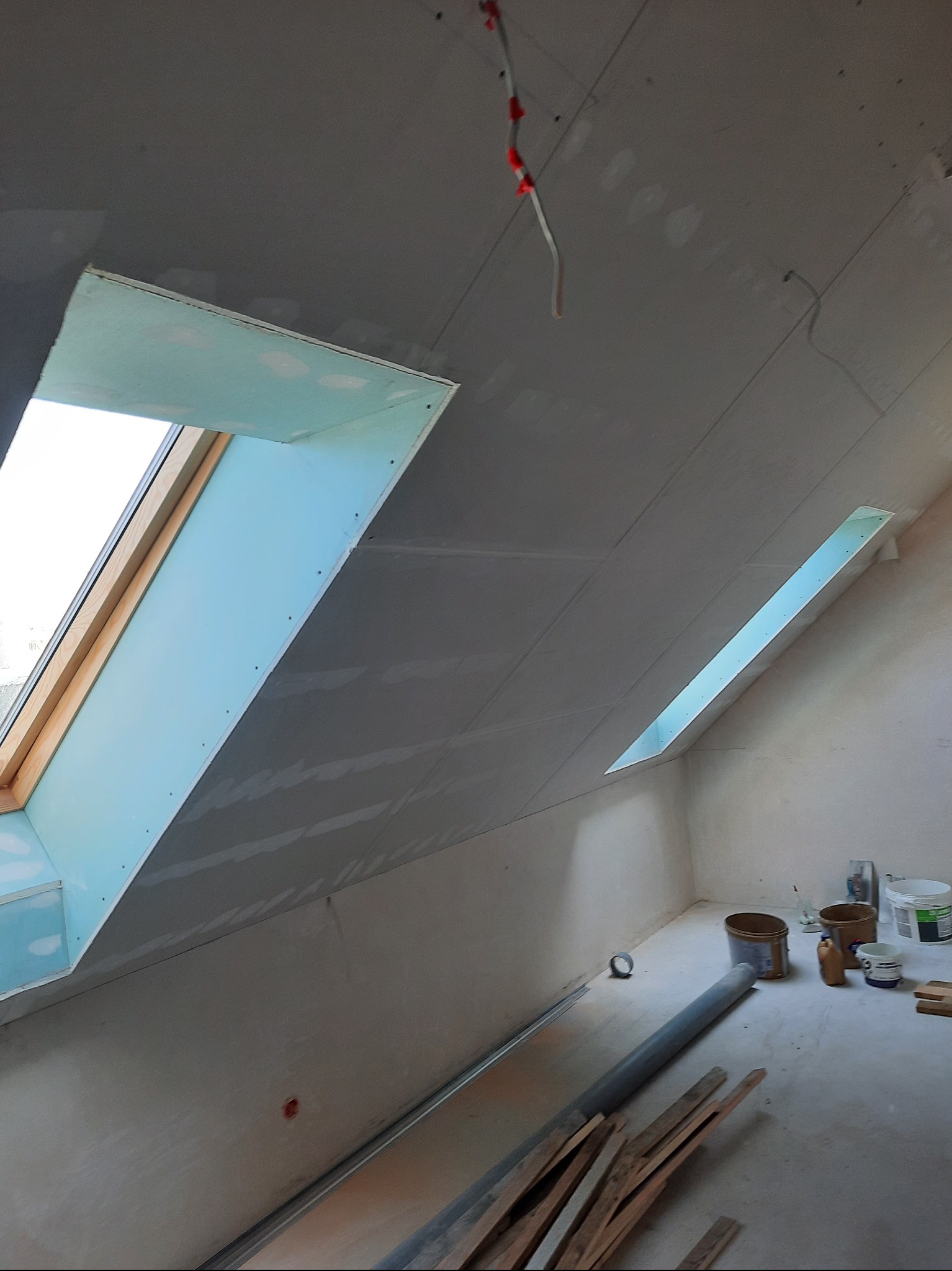 Poddasze z oknami dachowymi w trakcie montażu płyt gipsowo-kartonowych. Widoczne narzędzia i materiały budowlane. Skosy sufitowe przygotowane do dalszych prac.