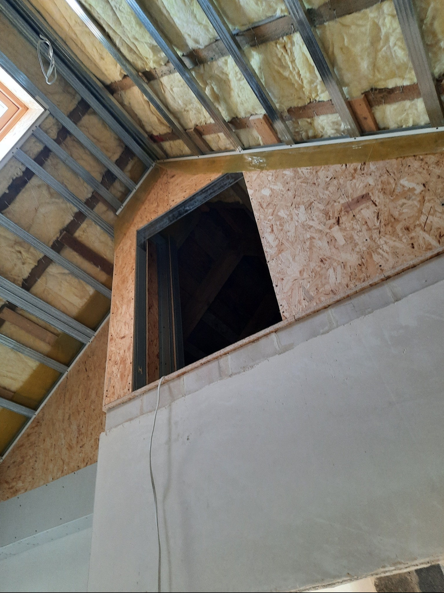 Skośne poddasze z izolacją, widoczne płyty OSB i metalowy stelaż. Fragment ściany z nałożoną gładzią. Okno dachowe w górnym rogu kadru.