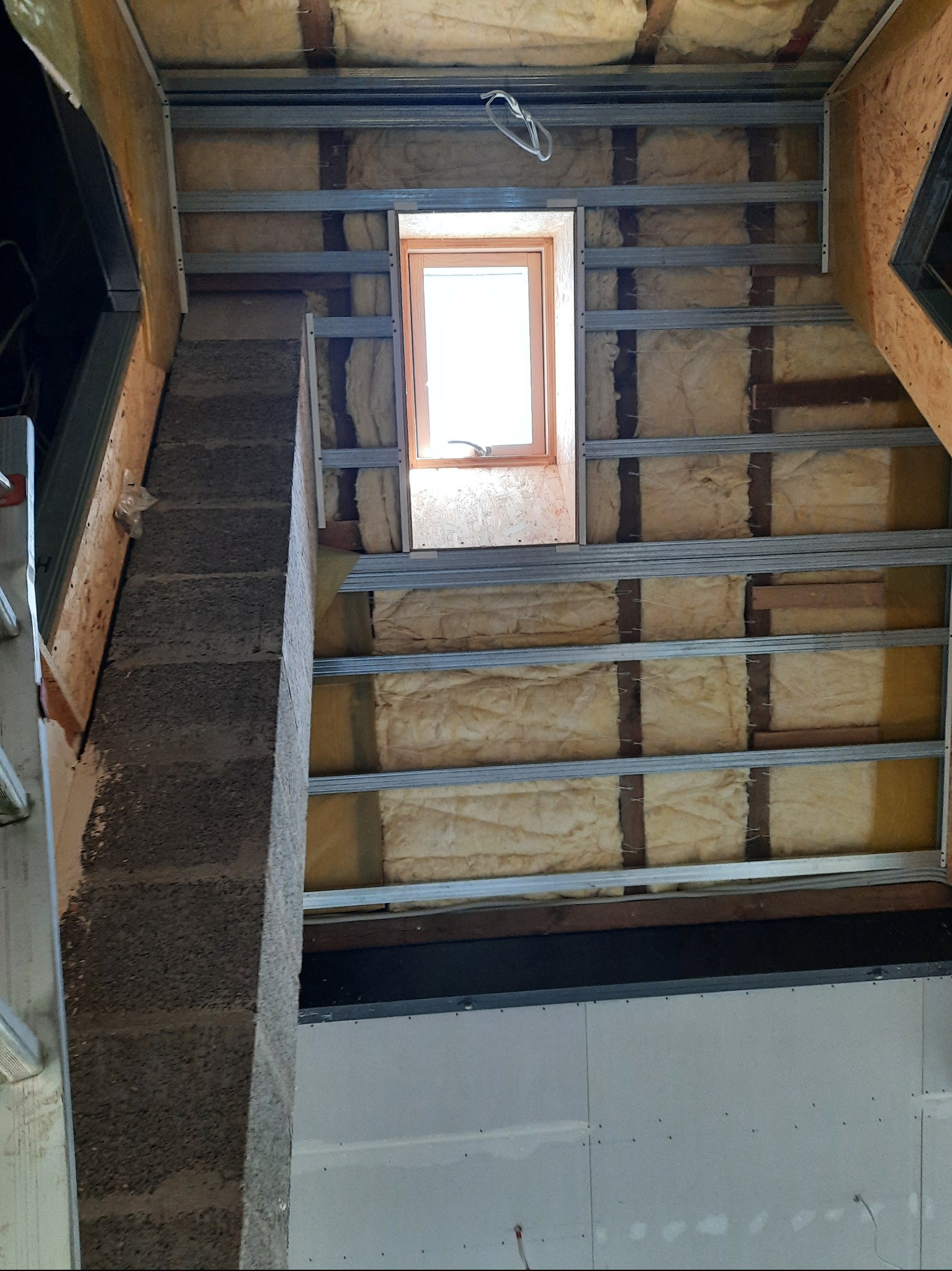 Poddasze z oknem dachowym w trakcie ocieplania wełną mineralną i montażu stelaża pod płyty G-K w Giżycku. Widoczny komin i fragment ściany z płyt.