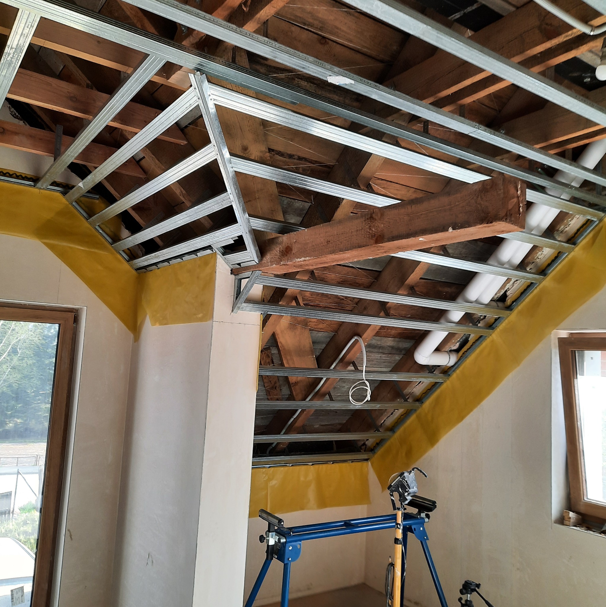 Montaż metalowej konstrukcji sufitu podwieszanego z widoczną izolacją i elementami instalacji. W tle drewniana więźba dachowa i okno.