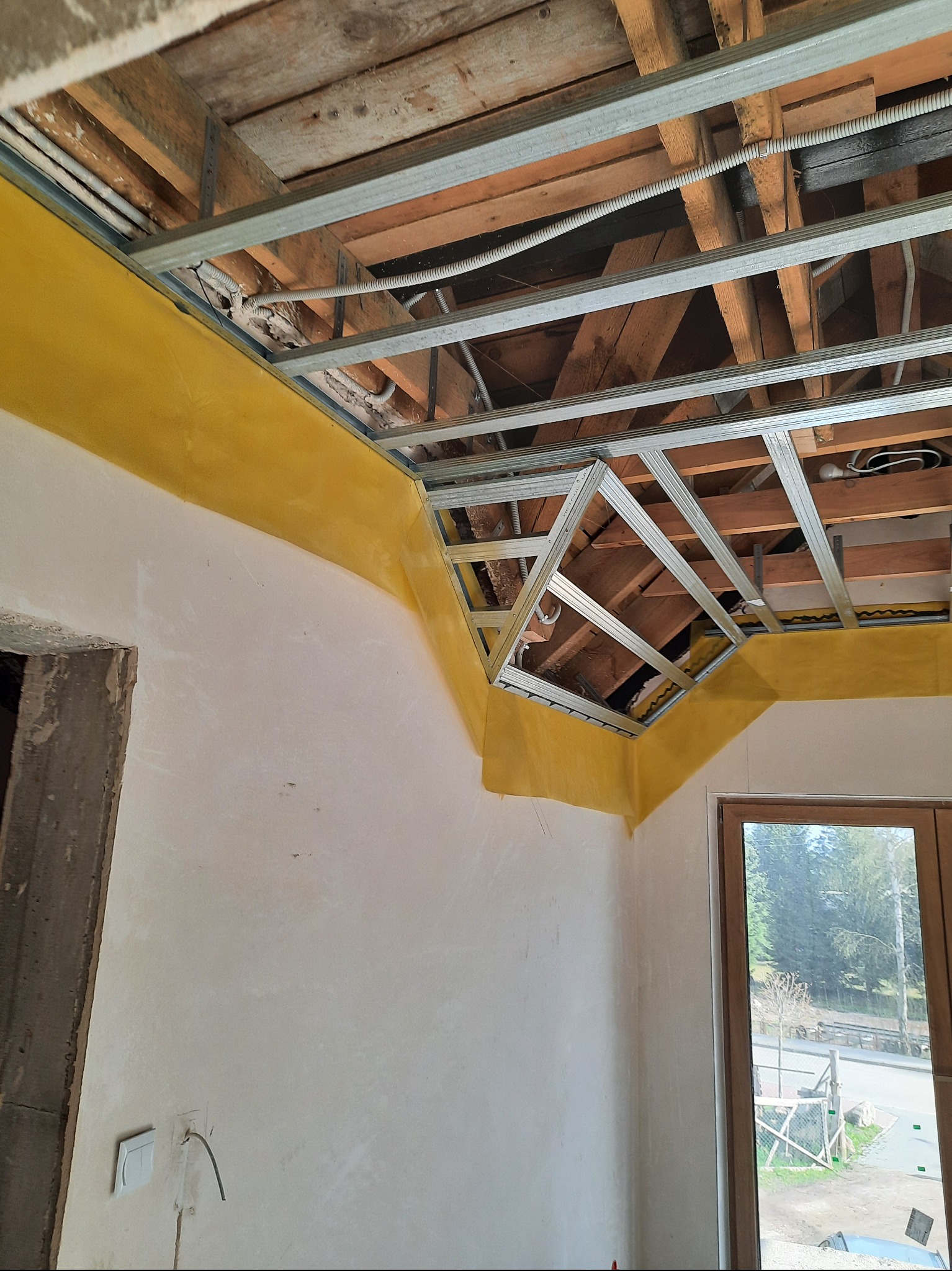 Sufit podwieszany: metalowa konstrukcja, żółta folia paroizolacyjna, drewniane belki stropowe. Widok na okno i surową ścianę.