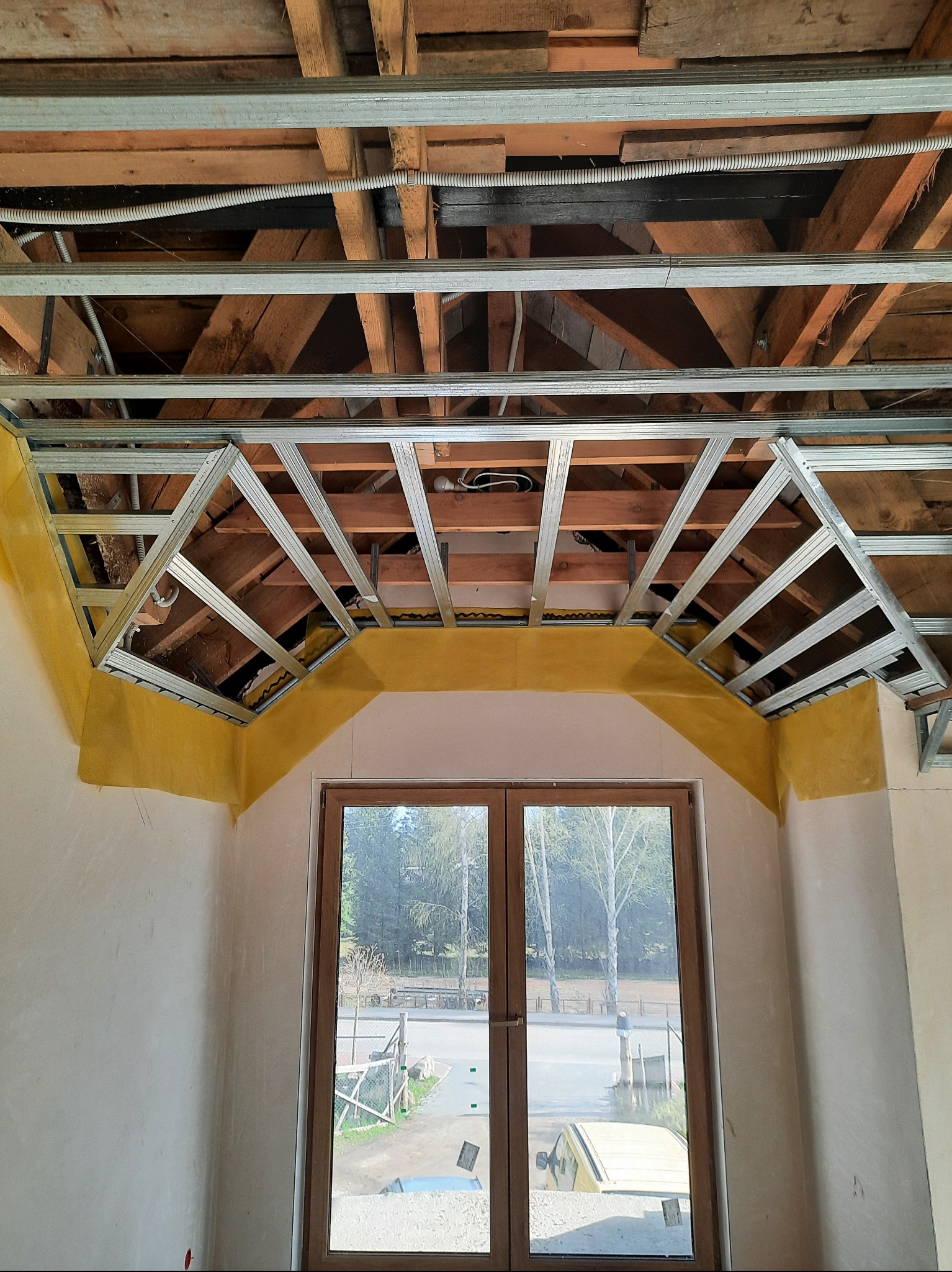 Sufit podwieszany z metalową konstrukcją i żółtą taśmą maskującą, widok z dołu na okno z widokiem na zewnątrz. Etap prac wykończeniowych.