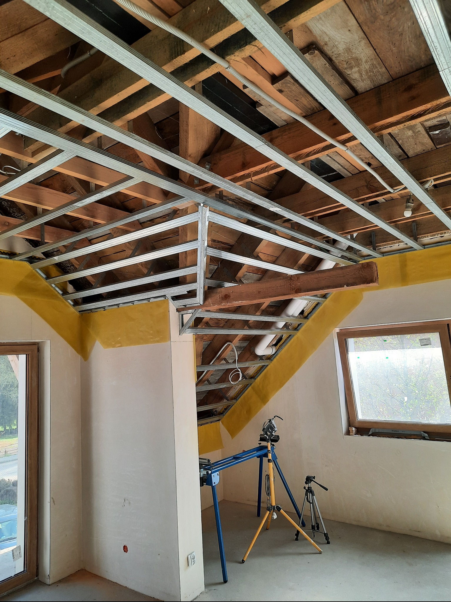 Montaż metalowej konstrukcji sufitu podwieszanego na drewnianym poddaszu, widoczne profile i żółta taśma uszczelniająca przy ścianach. W tle okno i narzędzia.