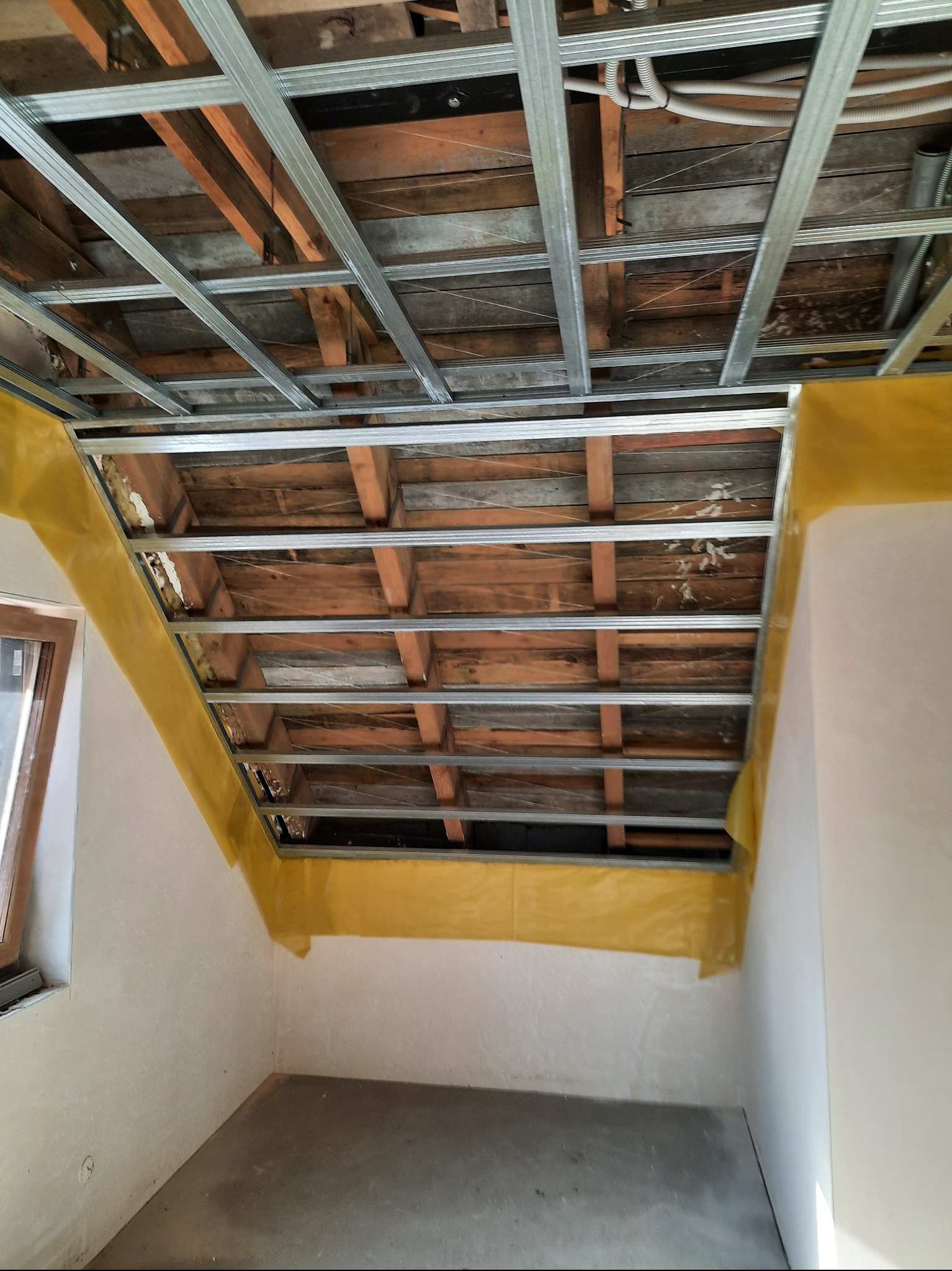 Skośne poddasze z metalową konstrukcją sufitu podwieszanego, zabezpieczone żółtą folią. Widoczne drewniane elementy konstrukcyjne i instalacje. Ściany przygotowane do dalszych prac.