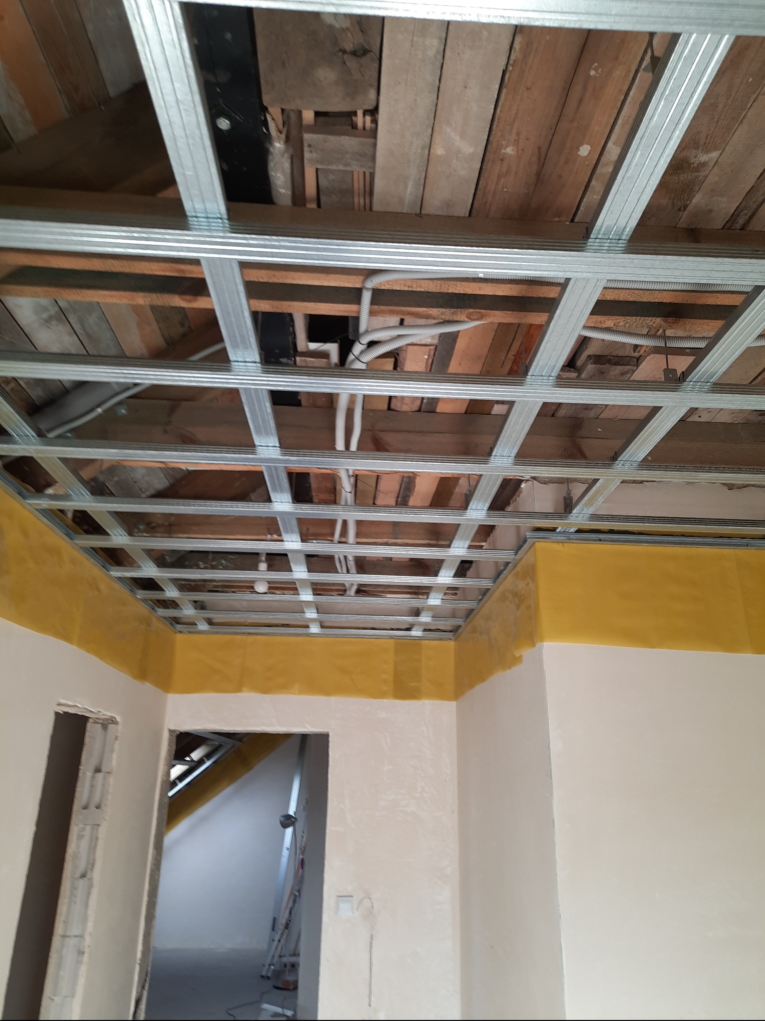 Metalowa konstrukcja sufitu podwieszanego z widocznymi instalacjami, częściowo zabezpieczona folią, w trakcie prac wykończeniowych wnętrza.