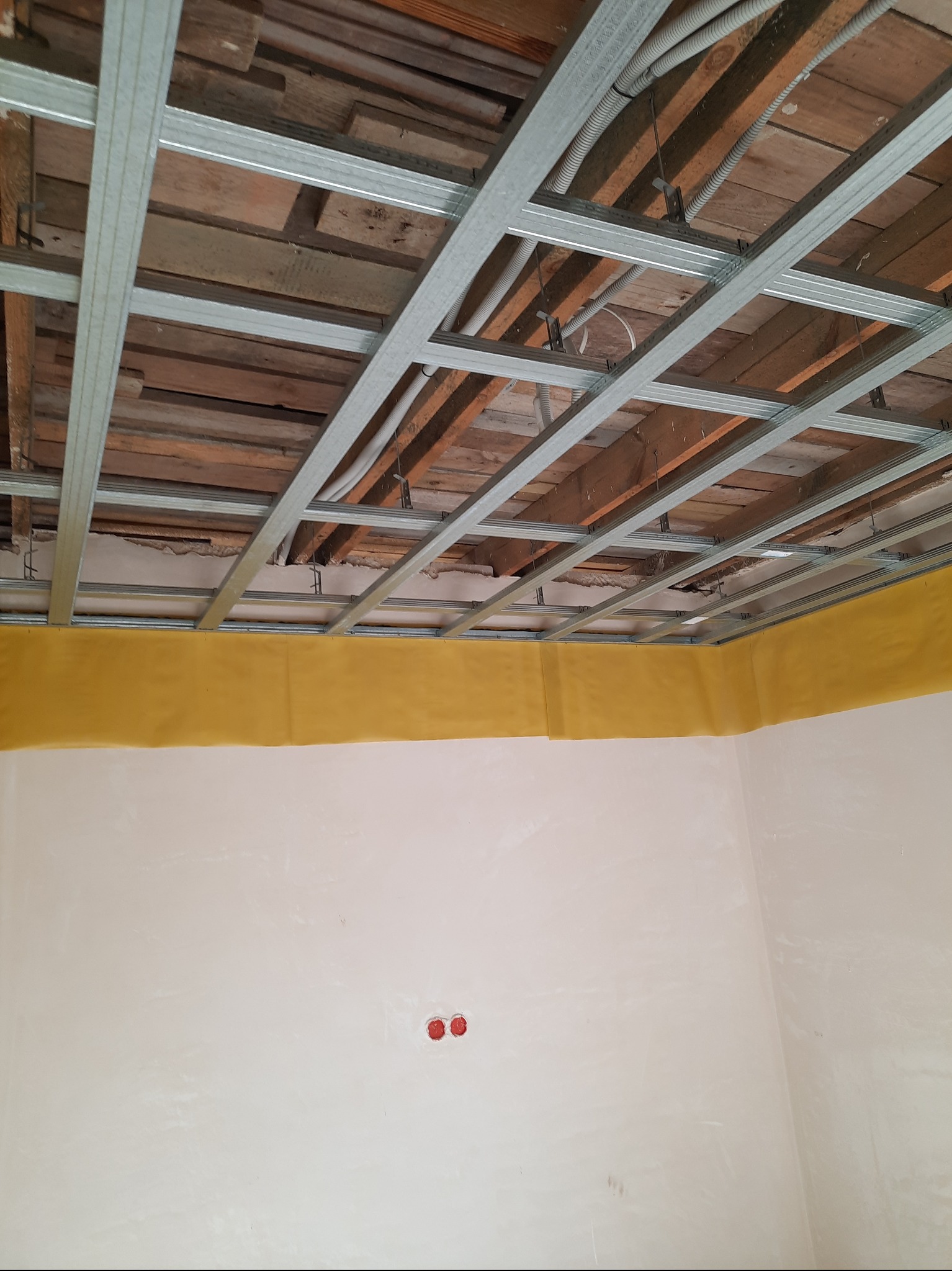 Sufit z metalową konstrukcją pod płyty gipsowe, widoczne drewniane belki stropowe i instalacje. Ściany przygotowane do dalszych prac wykończeniowych, z widocznymi puszkami elektrycznymi.