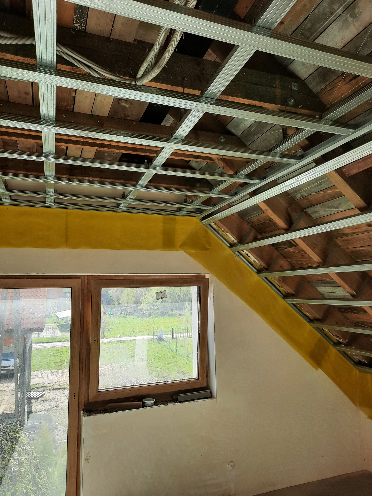 Szkielet sufitu podwieszanego z metalowych profili, widoczne drewniane elementy konstrukcyjne dachu, okno z widokiem na zewnątrz, fragment ściany z żółtą taśmą.