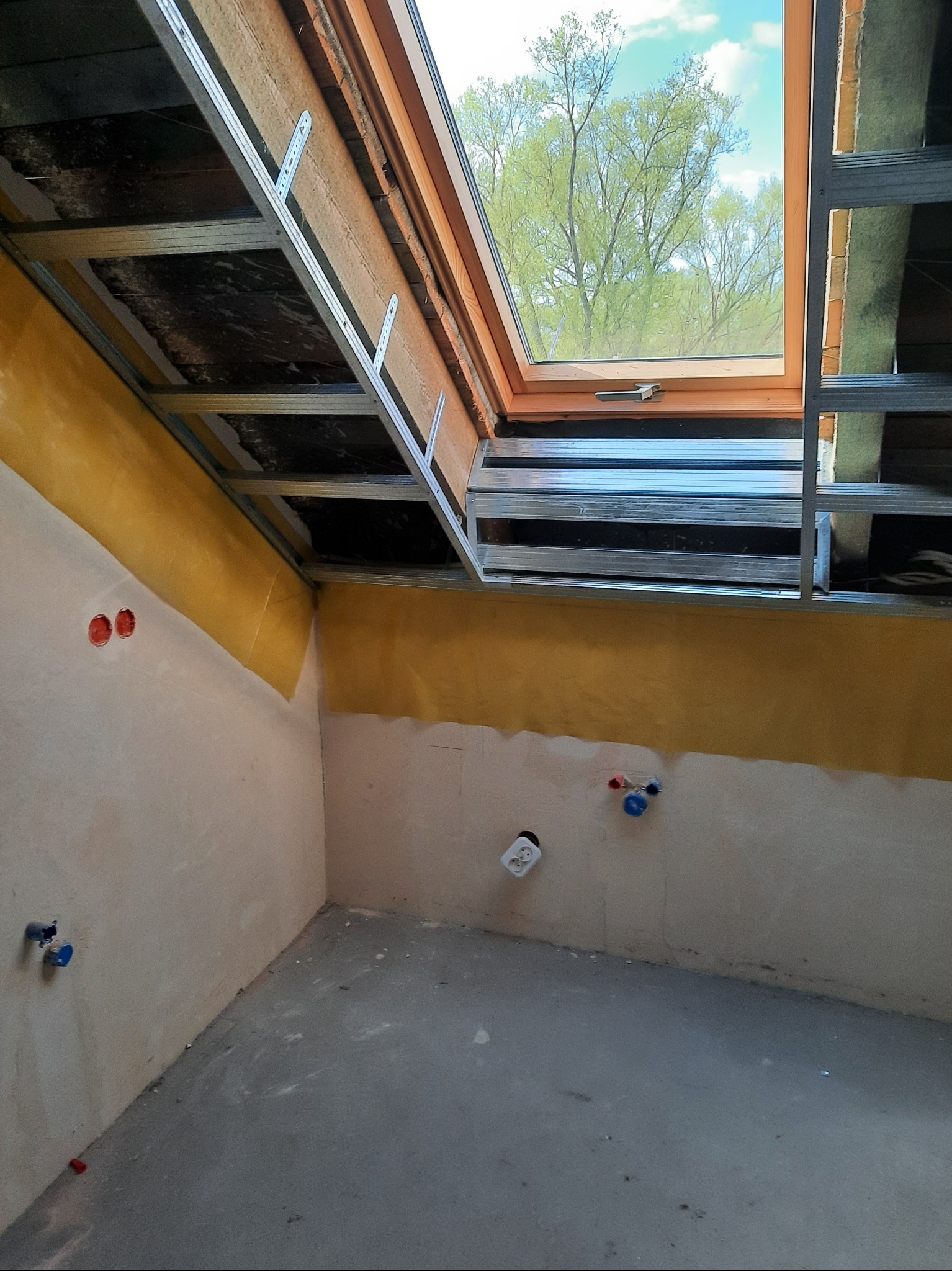 Poddasze w trakcie adaptacji: widoczne ocieplenie, metalowy stelaż pod płyty, okno dachowe z widokiem na drzewo, instalacje elektryczne i wodne na ścianach.