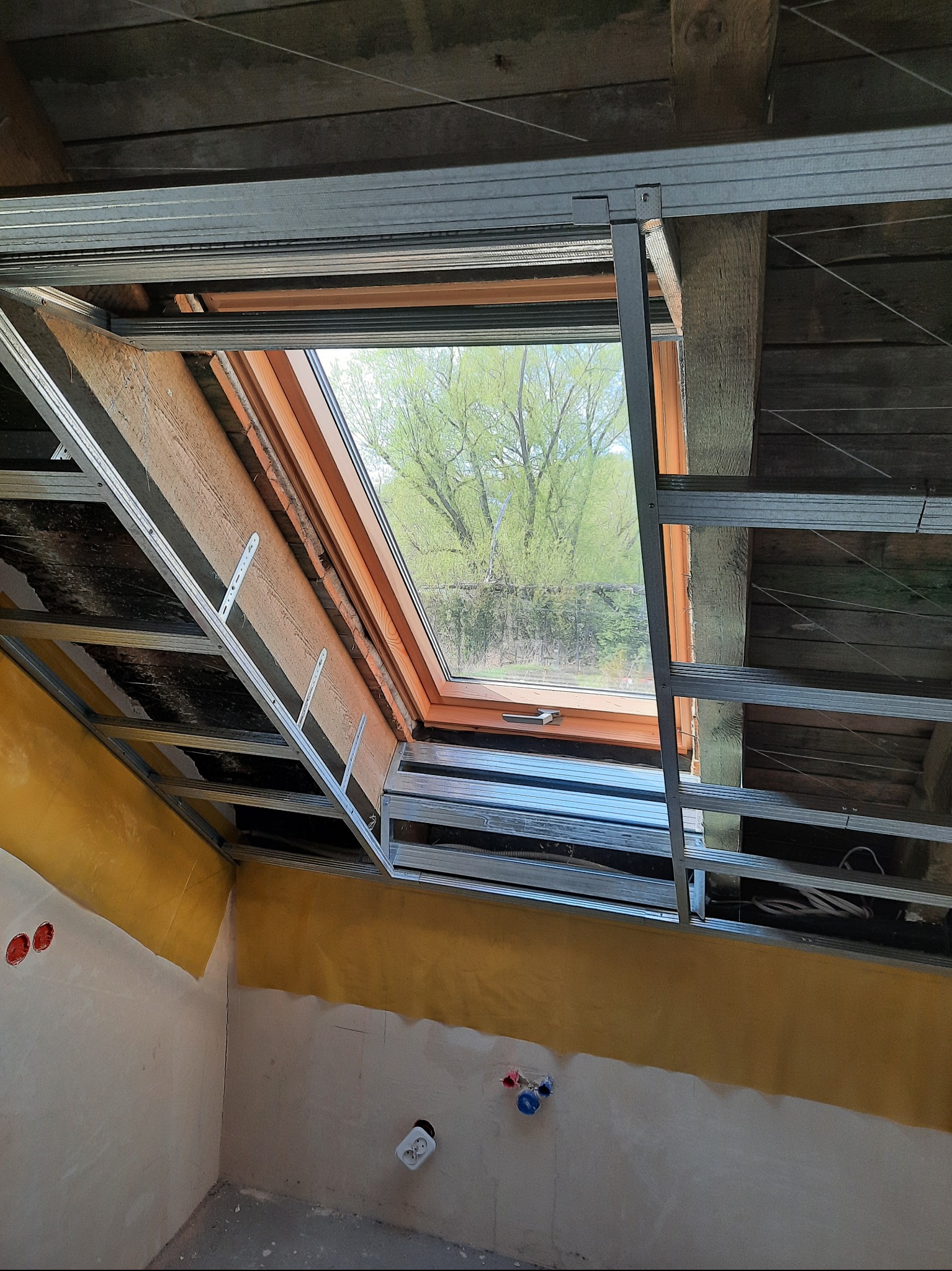 Montaż stelaża pod płytę karton-gips wokół okna dachowego na poddaszu. Widoczne drewniane elementy konstrukcyjne i fragment ściany z instalacją elektryczną i wodną.