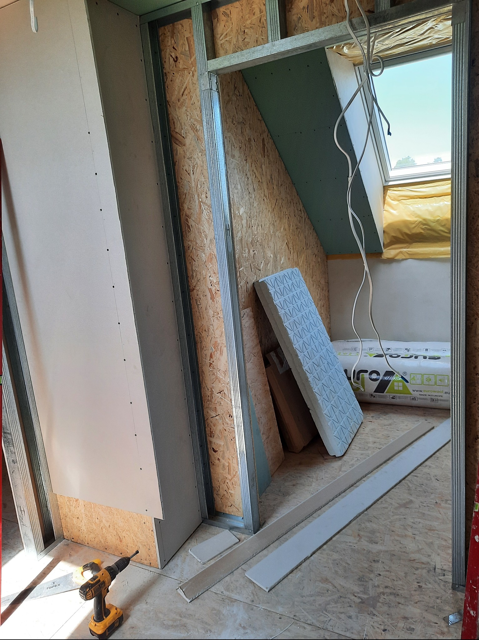 Skośne poddasze w trakcie zabudowy: metalowy stelaż, płyta OSB, częściowo zamontowana płyta gipsowa, okno dachowe, ocieplenie, wiertarka na podłodze.