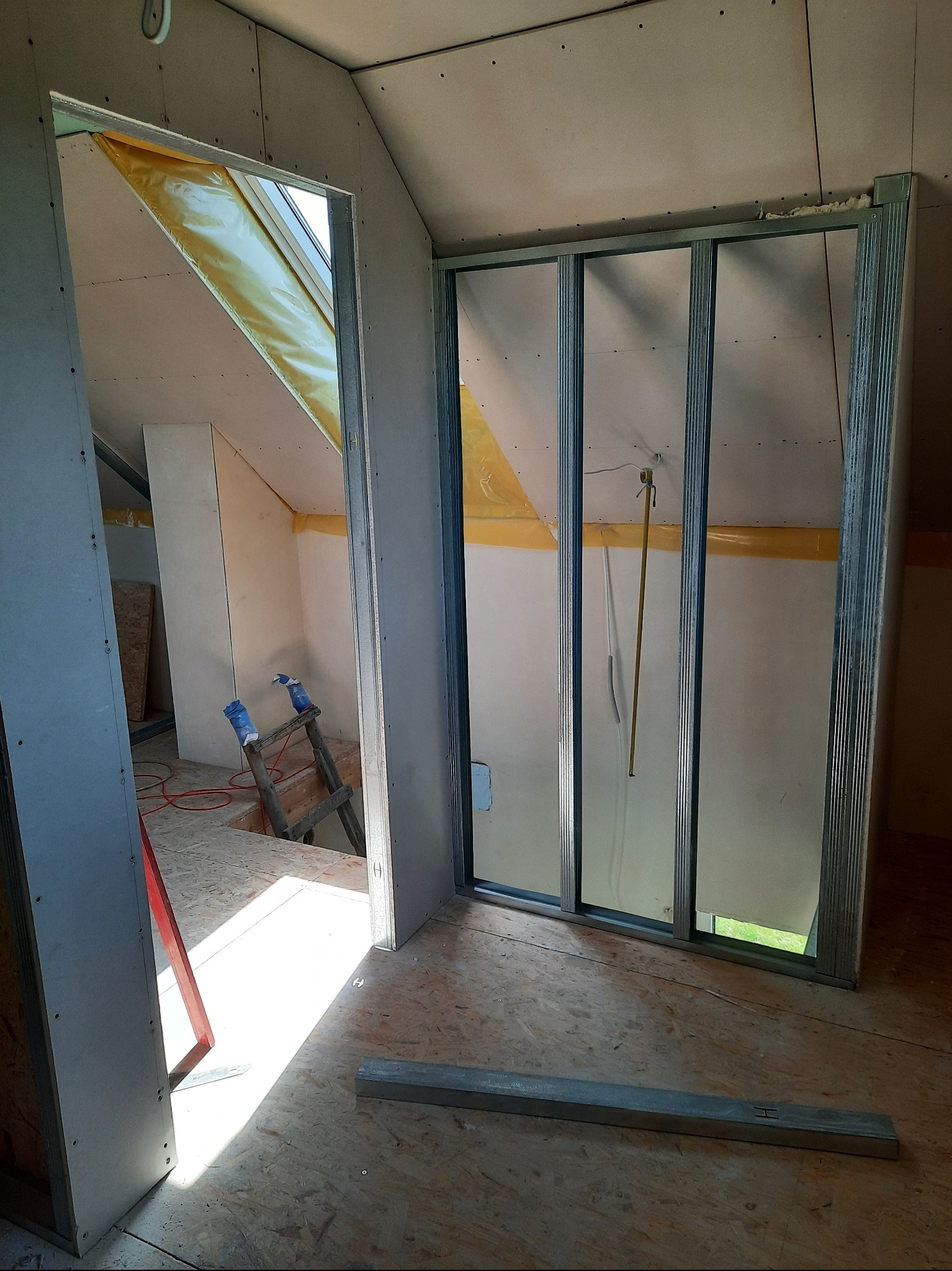 Poddasze w trakcie adaptacji: widoczne metalowe profile ścianek działowych, płyty gipsowe, ocieplenie okna dachowego. W tle drabina i narzędzia.