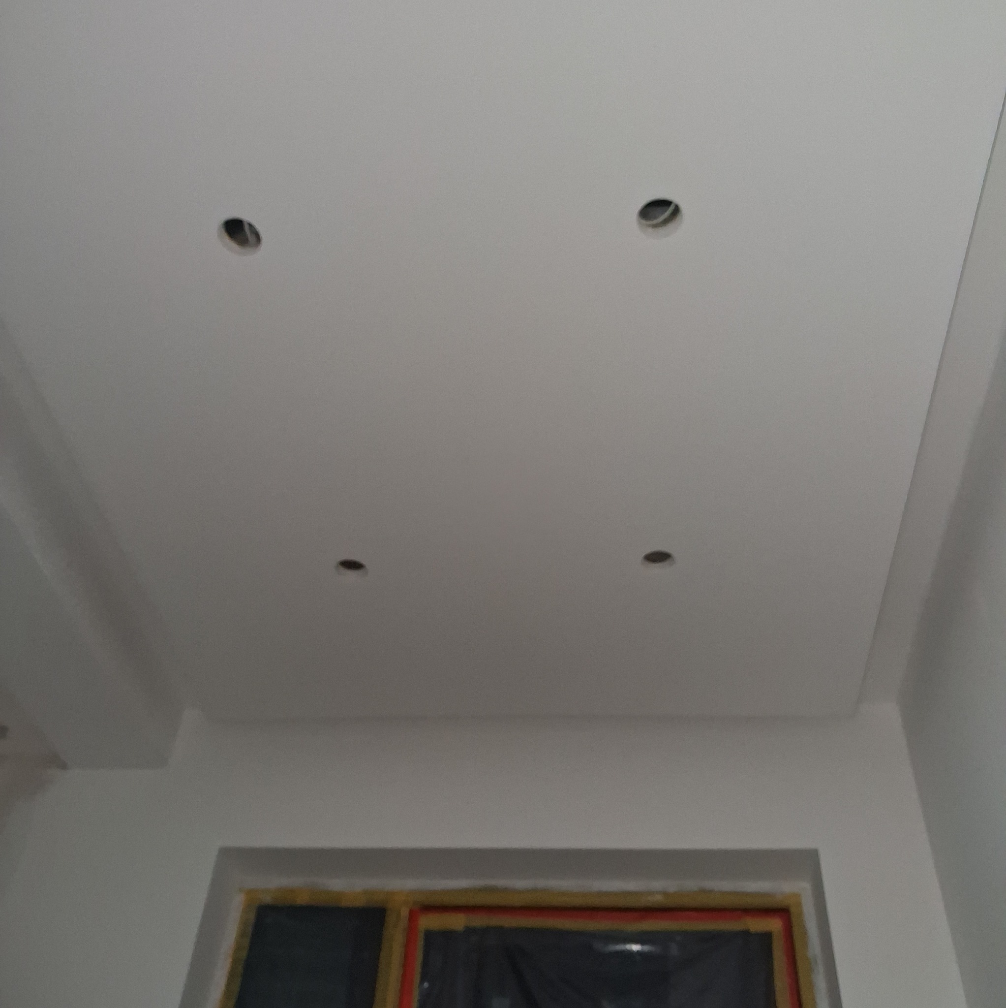 Gładki, pomalowany sufit z czterema otworami na oświetlenie punktowe, widoczne fragmenty ścian i okna zabezpieczonego folią malarską.